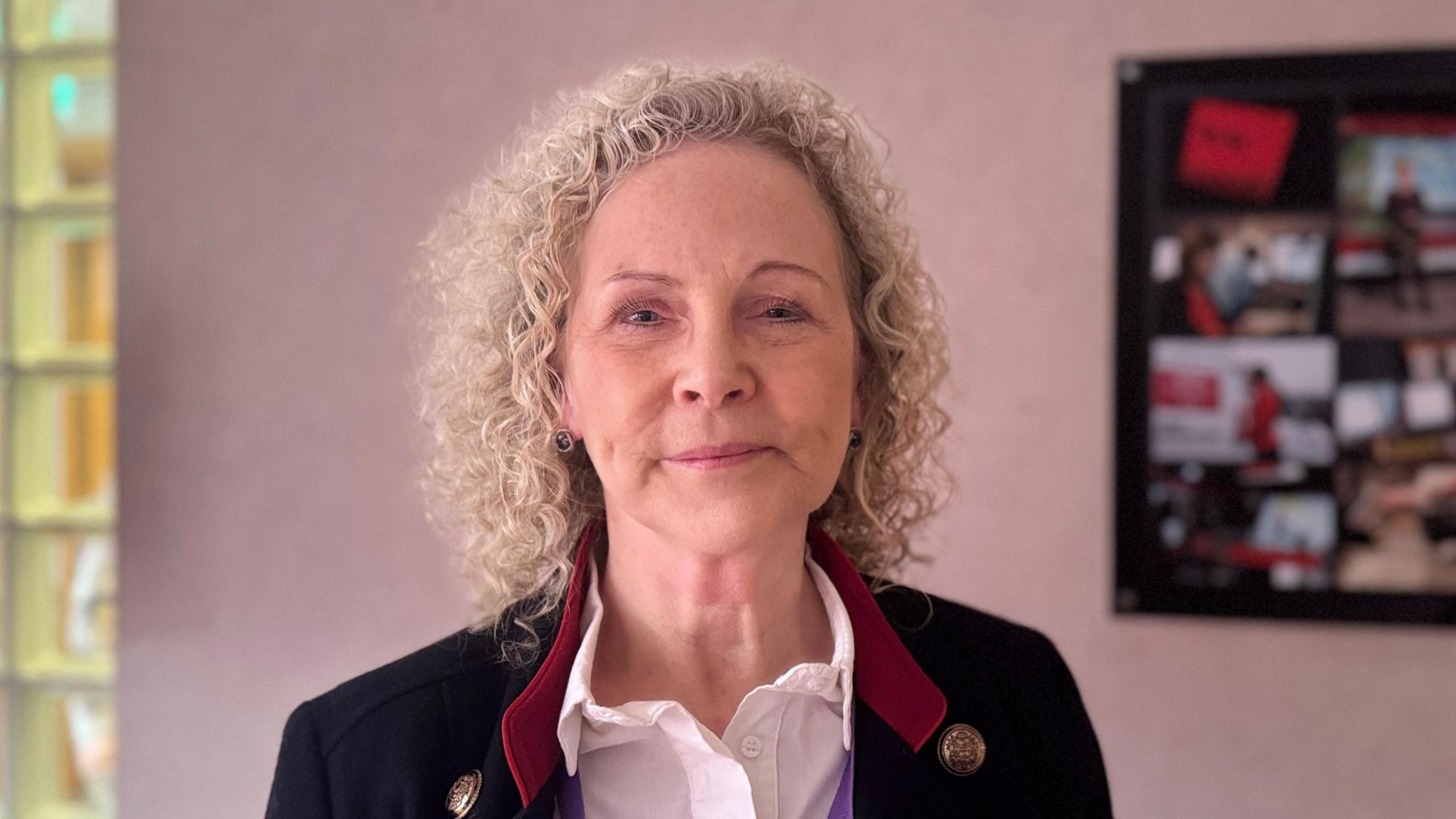 Dr Jacqui Montgomery-Devlin. She is an older woman with curly, blonde hair. She is looking at the camera with a neutral expression on her face. She is stood in a room with a pale pink wall, and a framed photo. She is wearing a white shirt, with a black blazer and red collar.