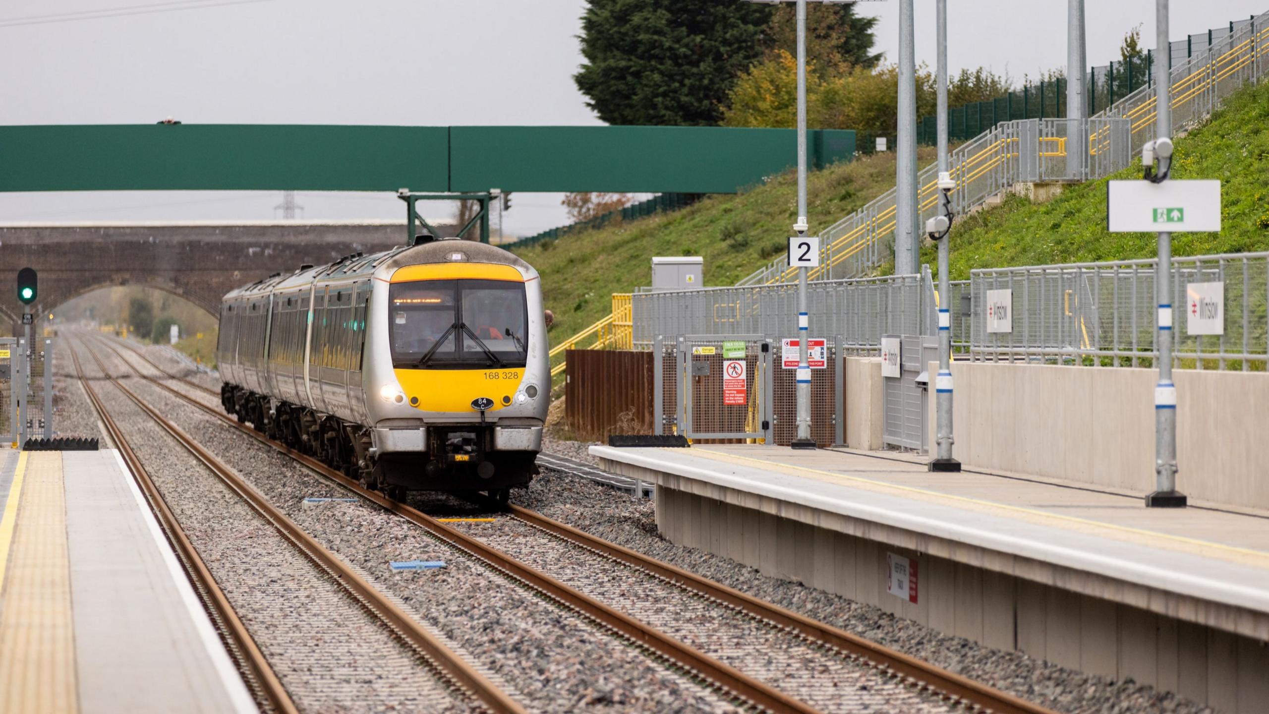 A yellow and grey train made up of three carriages is pulling into an empty platform two at a station on the Marston Vale line. The driver is wearing orange hi-vis and there is no destination listed at the front of the train.