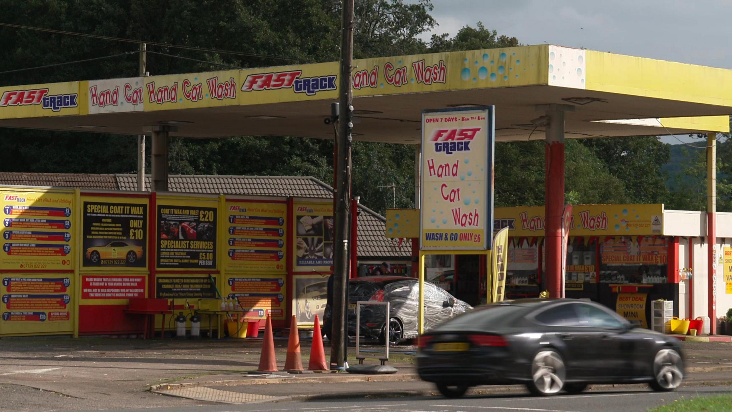 A black car is seen going past a hand car wash, which has yellow signage 