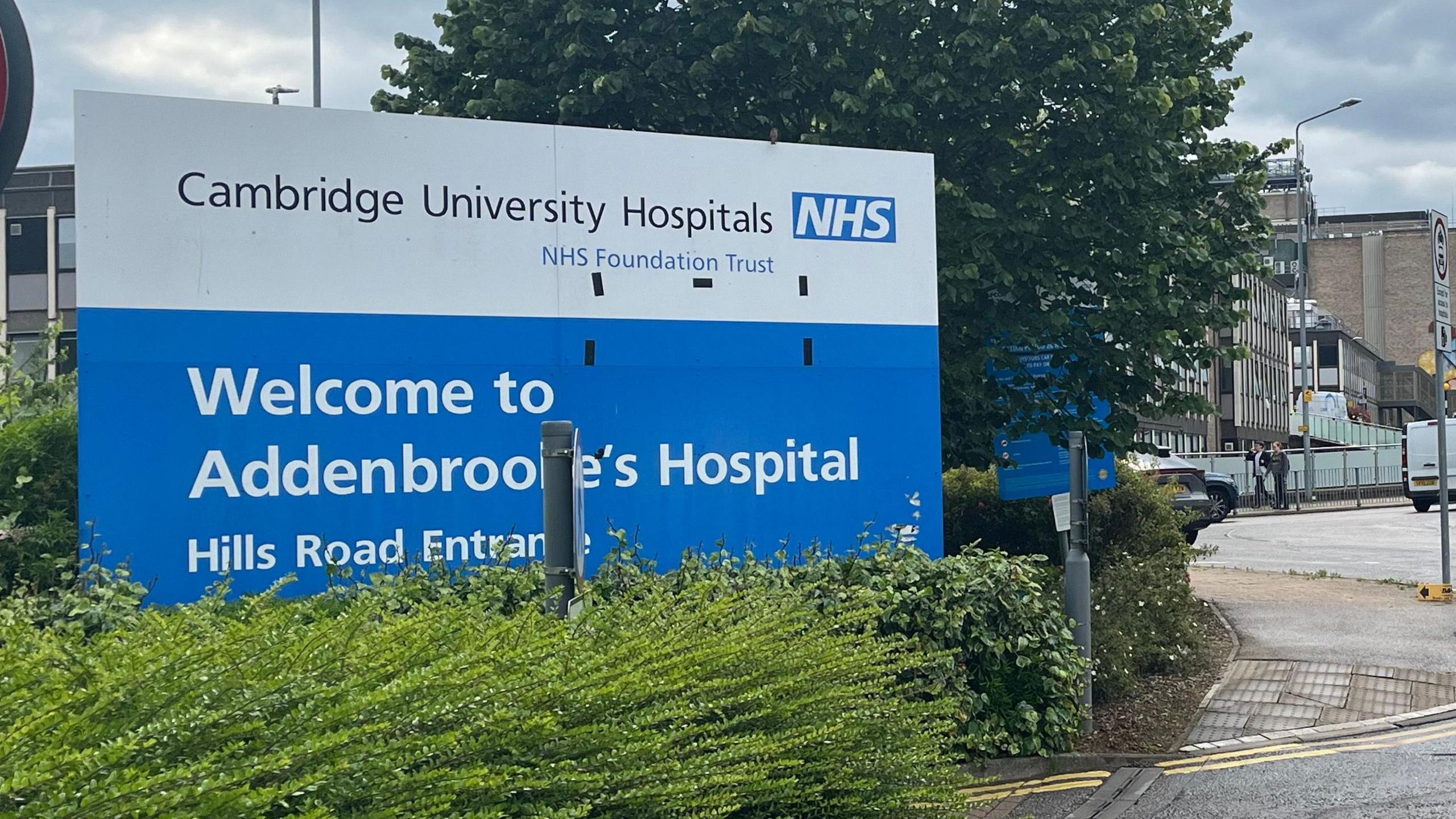 An NHS sign welcoming people to Addenbrooke's Hospital. It is blue and white and is printed with the words Cambridge University Hospitals NHS Foundation Trust. There are bushes, pavement, a road and buildings visible.