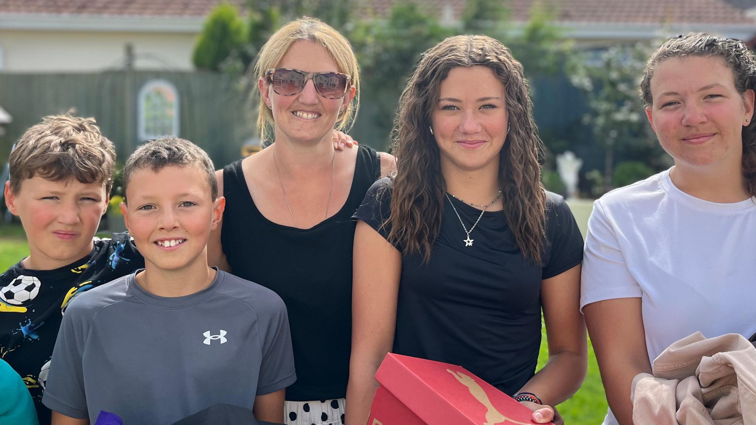 (left to right James (10) , Henry (11), Mum Nikki, Grace (14) and Olivia (16)). They are all smiling holding their back to school purchases and hand-me downs. The family is standing in the garden on a sunny day. They all have black hair apart from the mum who is blonde.