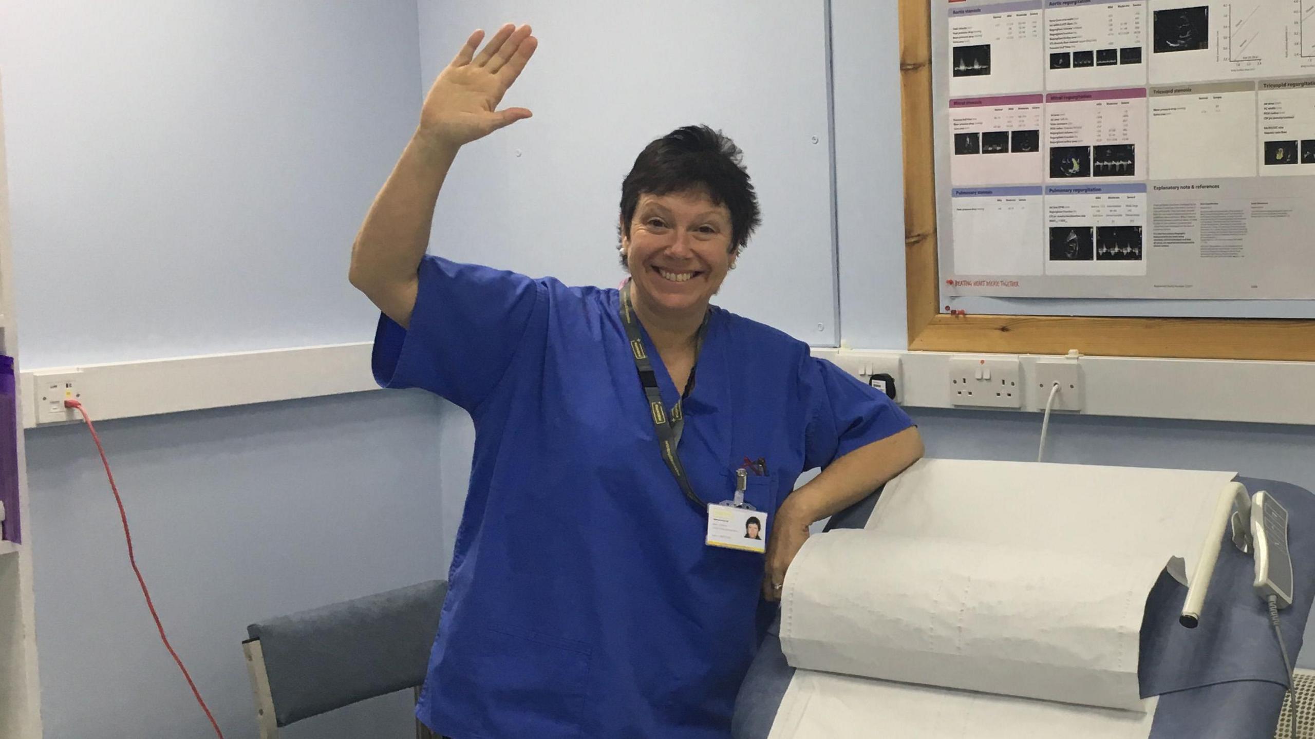 Jane Graham, wearing dark blue hospital scrubs and a lanyard, waves in a hospital consultation room