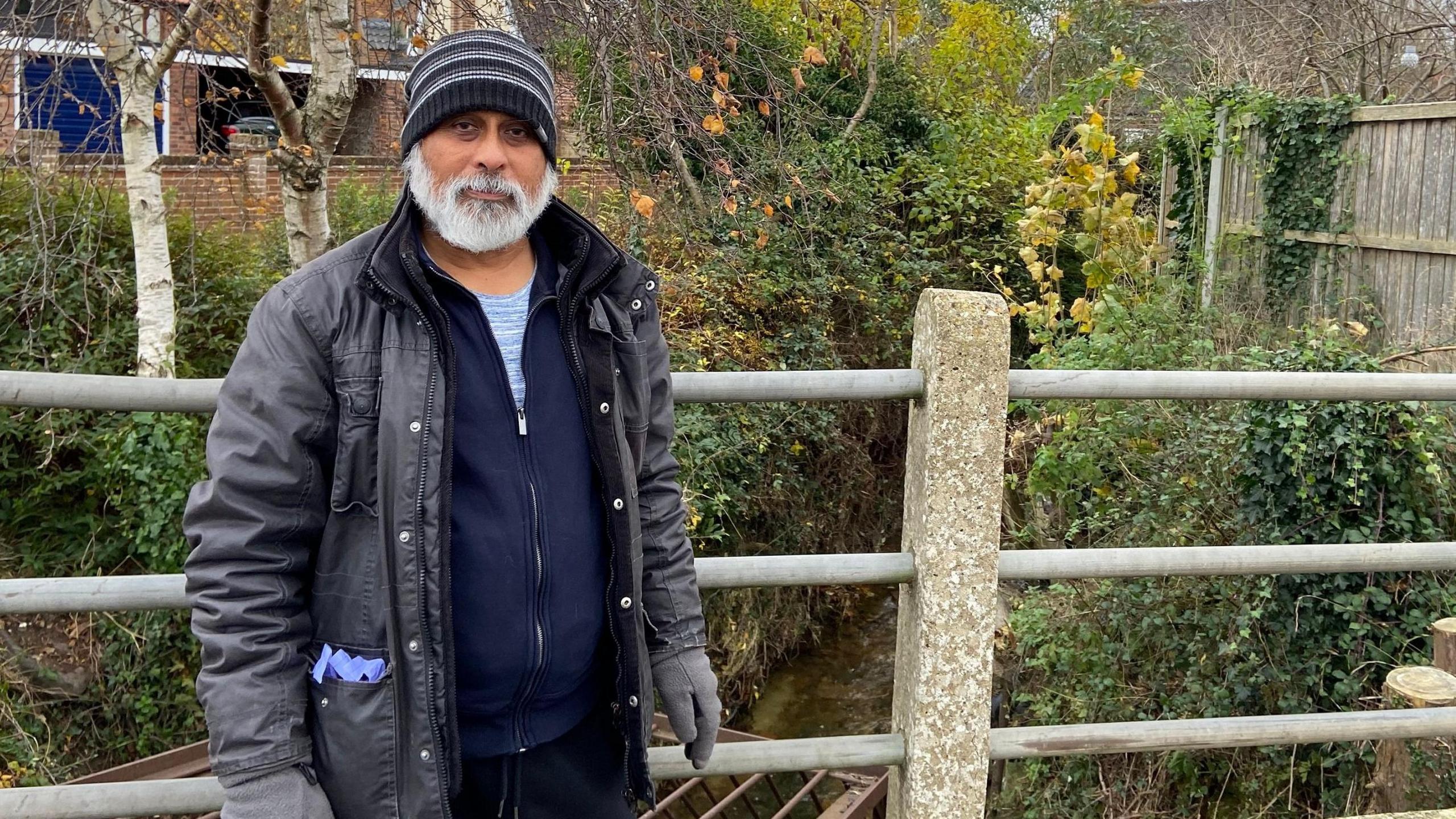 A man with a big white beard and dressed in a coat and gloves and hat is stood near a river on an autumnal day