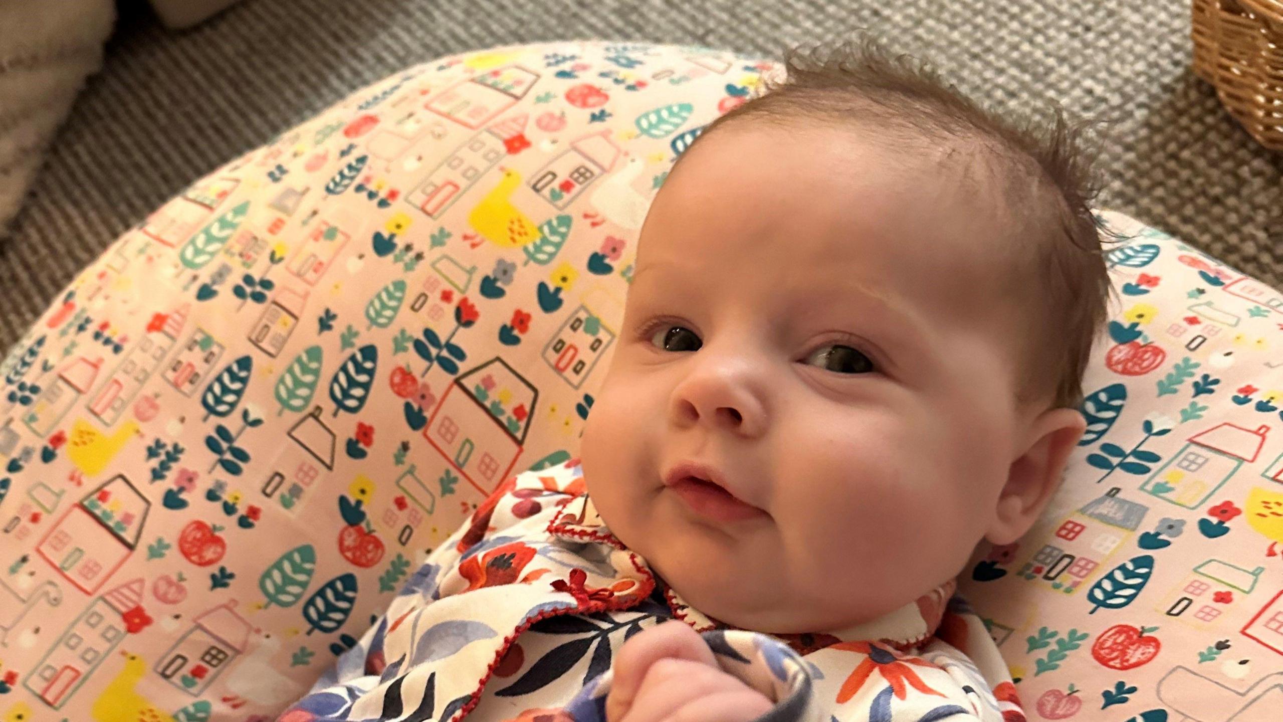 A baby with a small amount of brown hair wearing a floral patterned onesie lying on a pink cushion.