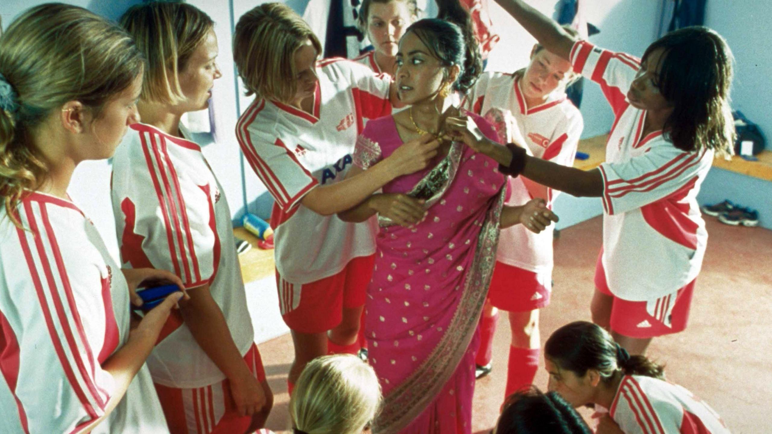 A scene from Bend it Like Beckham in which a group of white girls in football kit fit a pink sari with gold embroidery on to an Indian teenage girl, Parminder Nagra