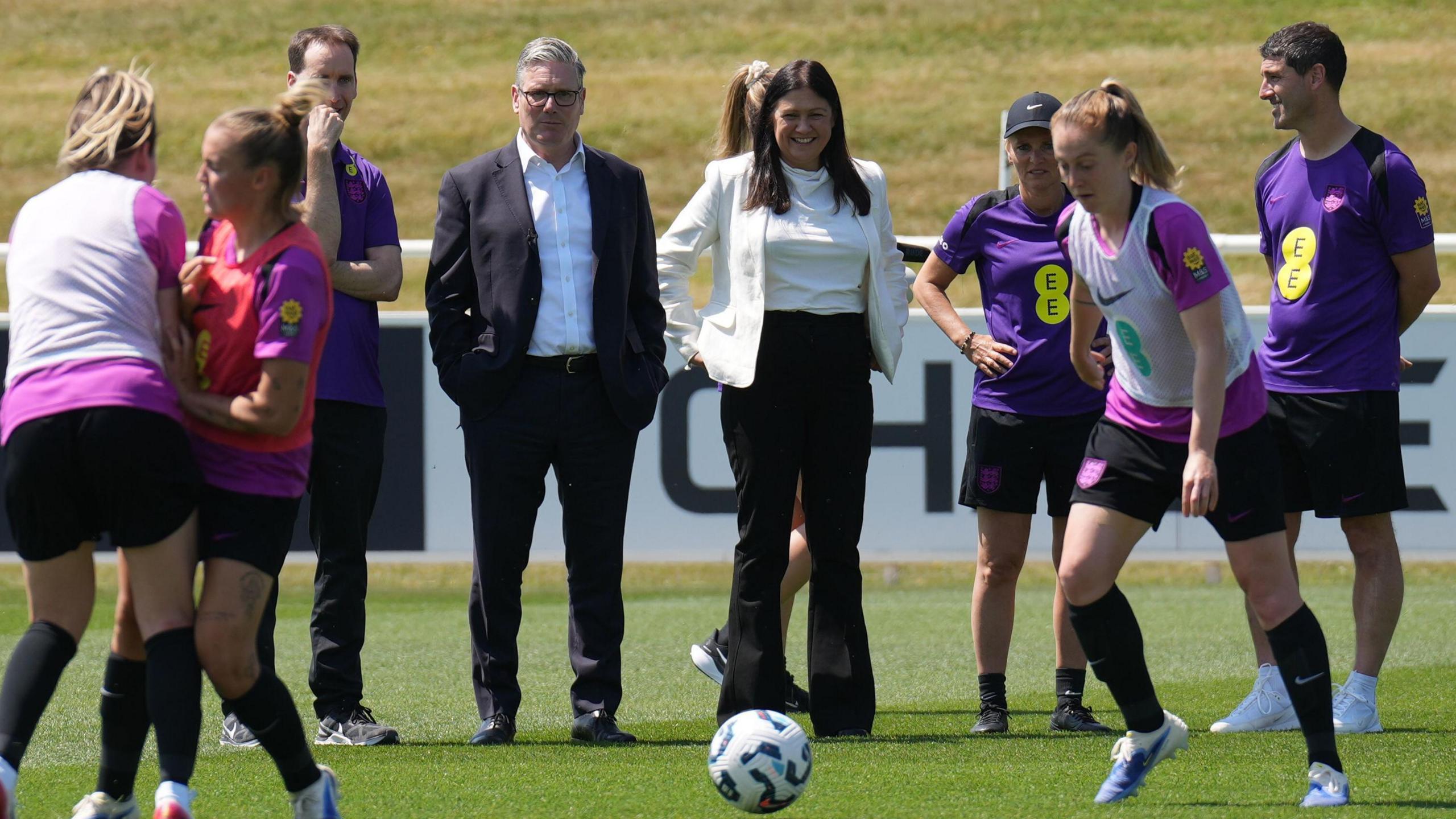 The Prime Minister, Sir Keir Starmer and The Secretary of State for Culture, Media and Sport Lisa Nandy, both dressed in suits, watch and England training session on a green pitch.