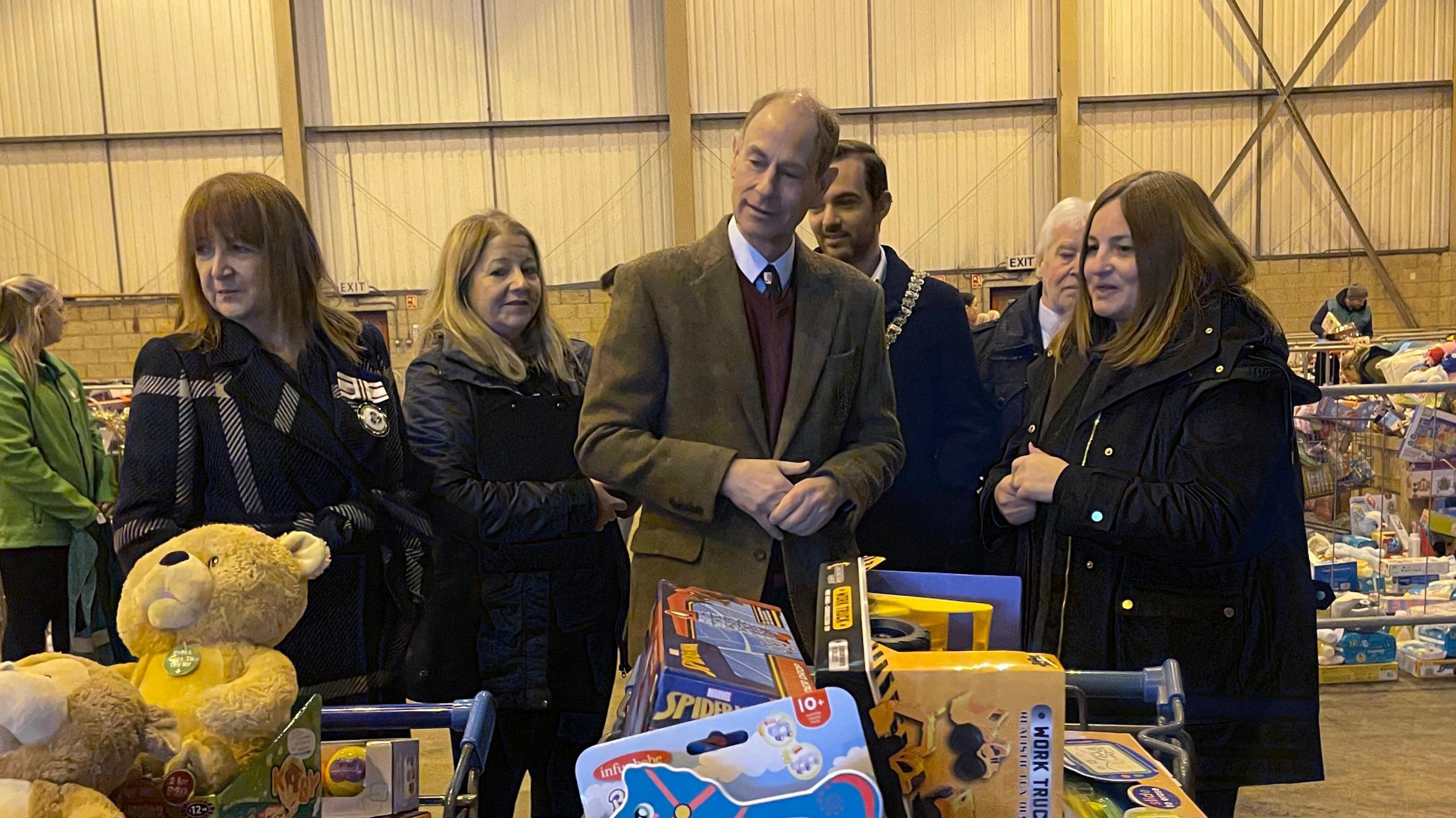 The Duke of Edinburgh stands amongst five people in front of trolleys of children's' toys. He is wearing a green blazer, burgundy sweater and white shirt. Those around  him are wearing dark coloured coats.