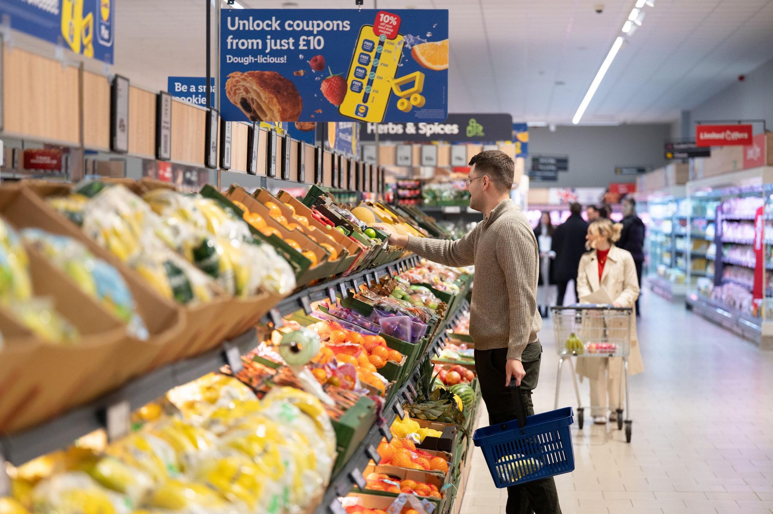 A man wearing dark brown trousers and a light brown jumper in the fruit and veg aisle of Lidl carrying a basket and reaching for the fruit