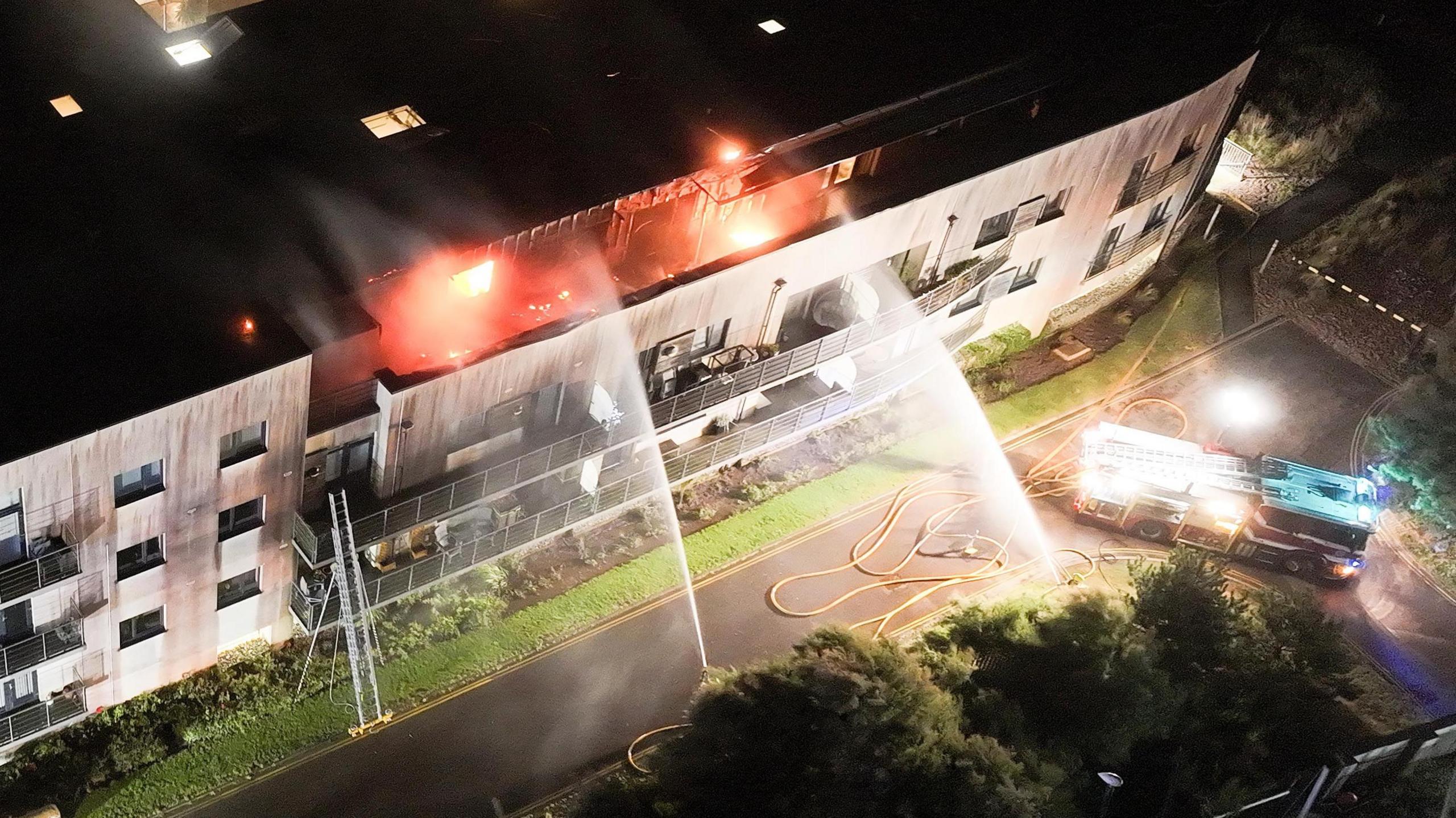 An aerial shot of water being sprayed towards the top floor of a flat with a fire engine at the base of the building