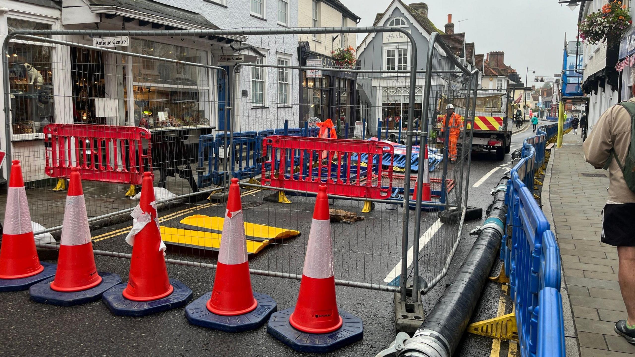 A section of road which has been closed off with red and white cones and metal and red plastic barriers. A pipe runs along next to the pavement, with blue barriers to the right of it. Workers in hi-vis uniforms and a flatbed truck are in the background