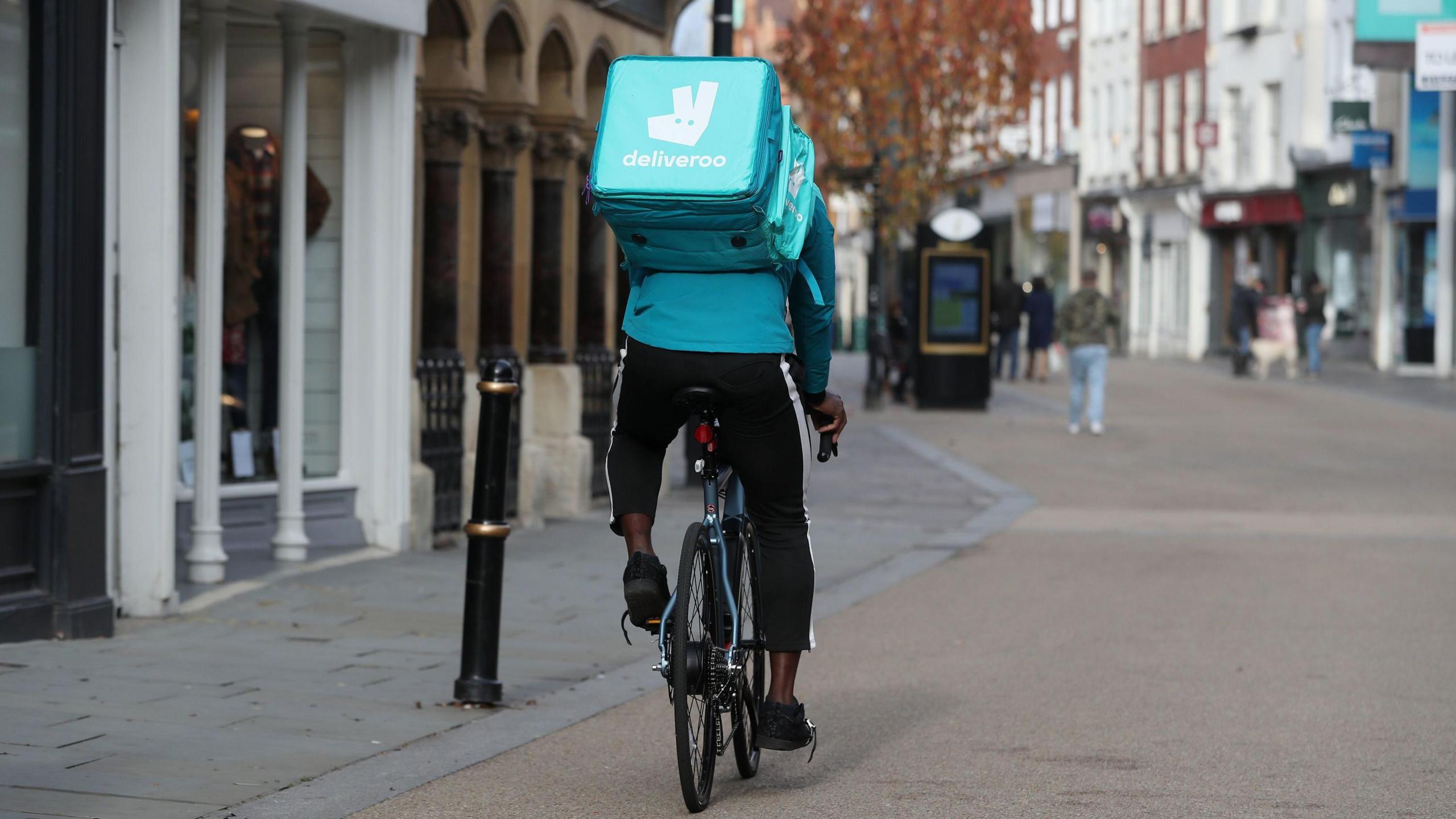 Deliveroo rider on bike wearing black trousers, bright blue top, and bright blue square Deliveroo bag on his back saying the firm's name and with its rabbit logo on it