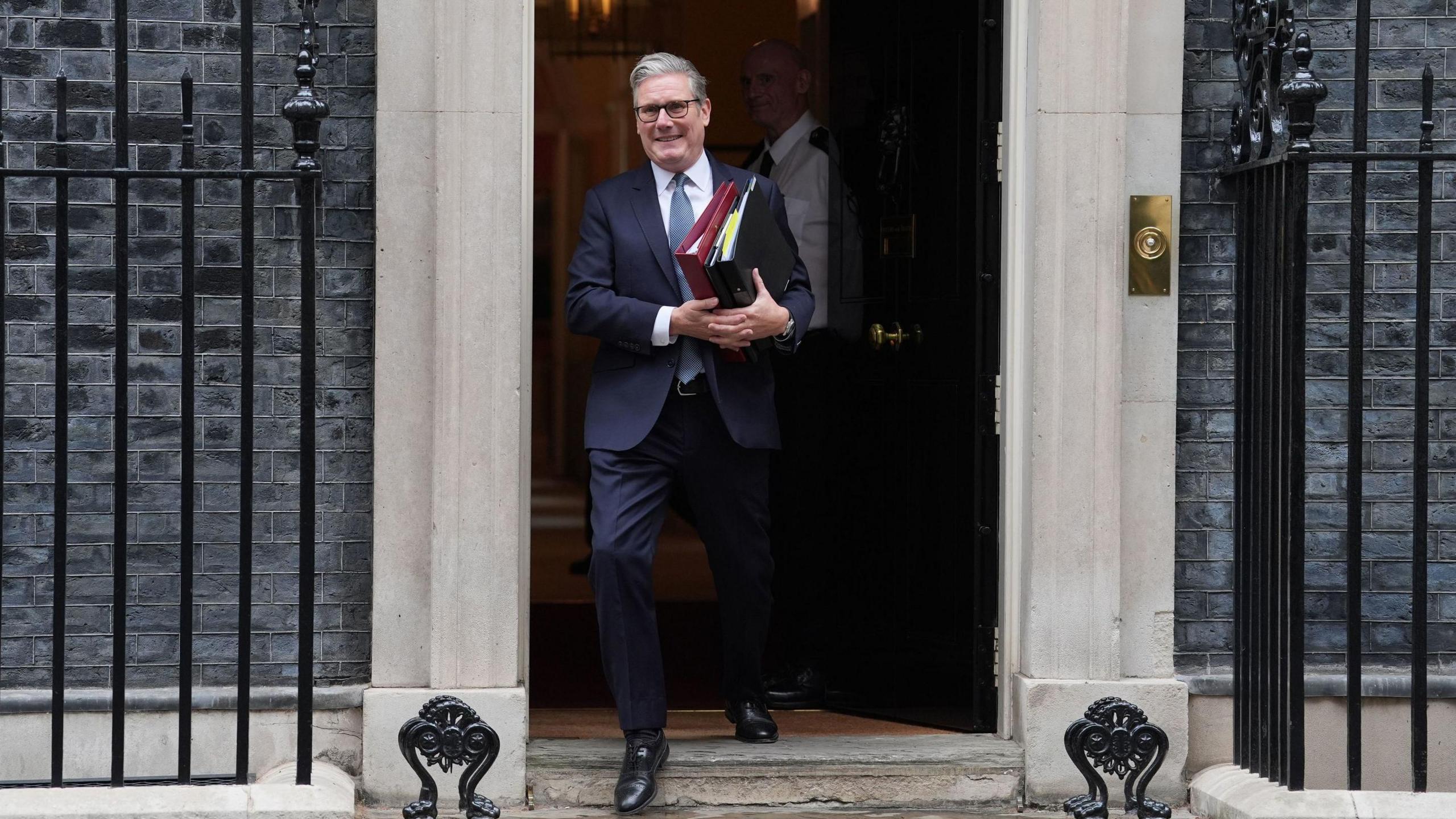 Sir Keir Starmer leaving 10 Downing Street ahead of PMQs - he is carrying folders and is dressed in a navy suit with a blue tie. He has grey hair and is wearing dark-rimmed glasses.