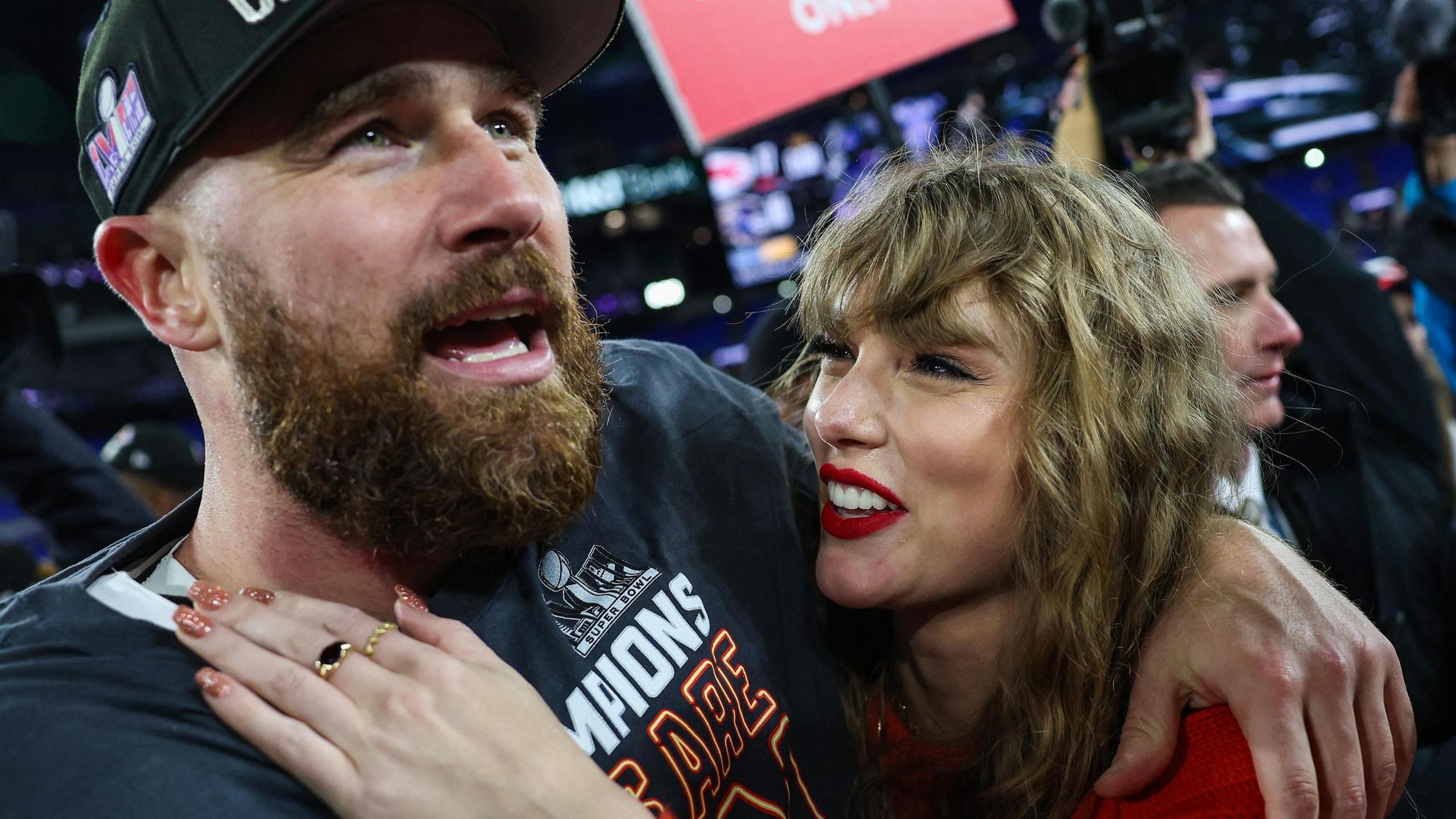 Travis Kelce and Taylor Swift with their arms around each other and her smiling and looking at him