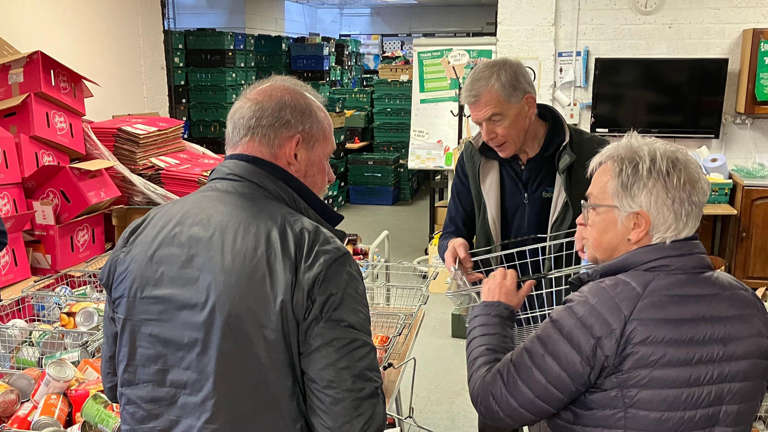 Two men and a woman look at some of the shopping baskets which have not yet been filled with foodstuff.  All three volunteers are wearing grey type jackets and fleeces underneath.