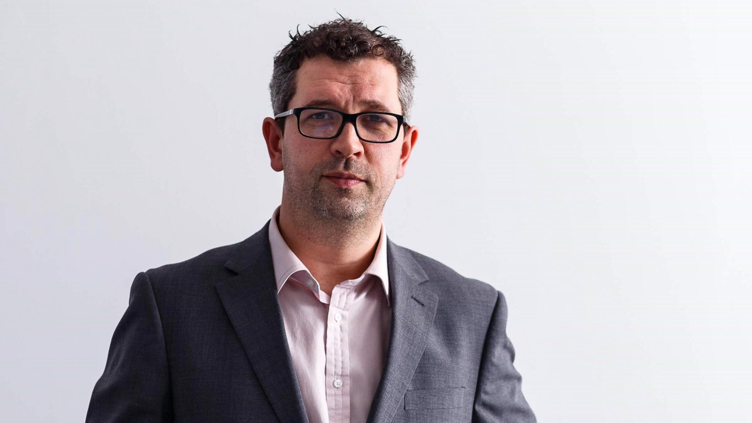 A professional headshot of Dan Panes. He stands against a white backdrop and wears a grey suit jacket and a white shirt. He has short dark brown hair and wears black rimmed glasses.