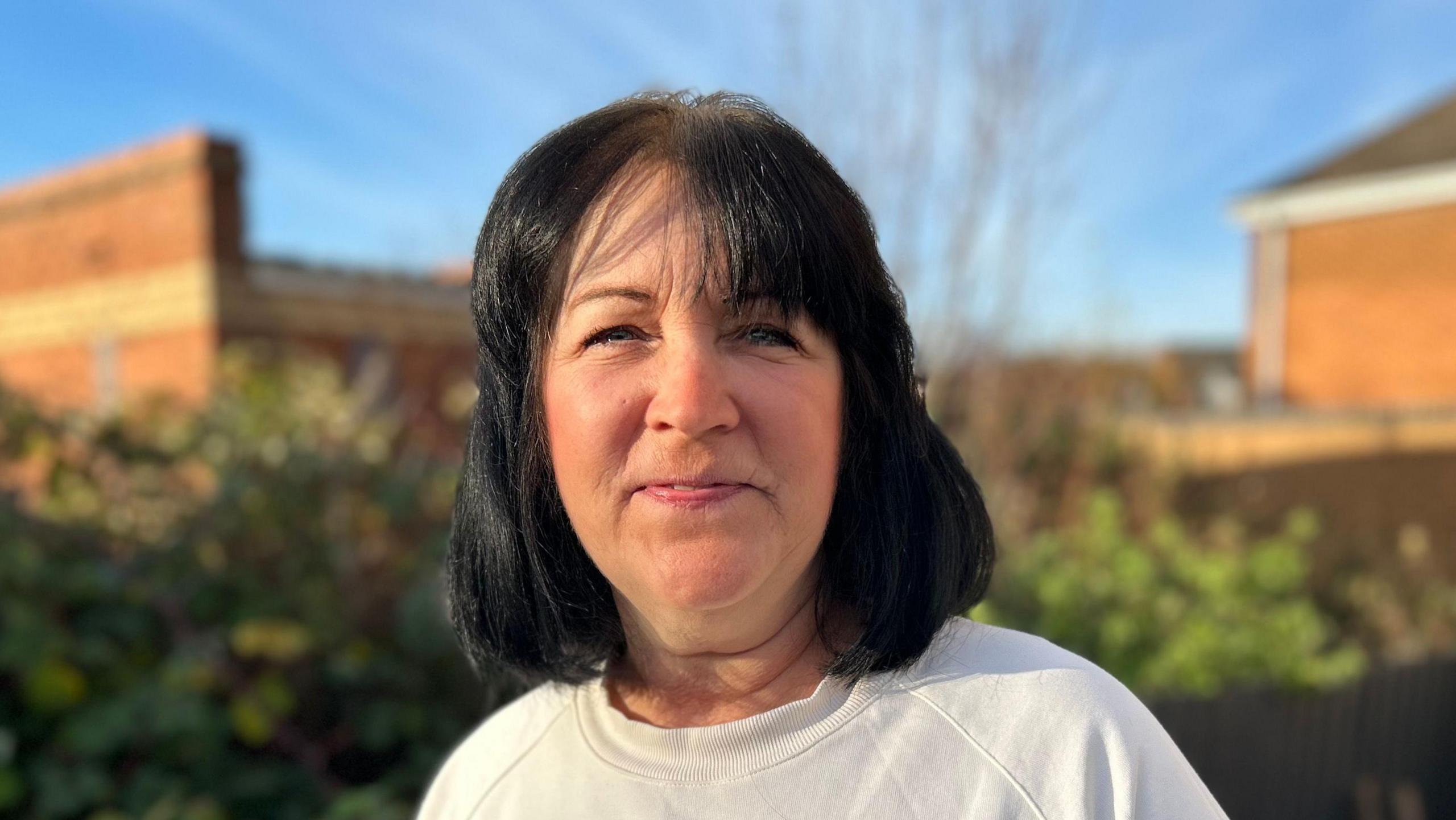 A woman with shoulder length black hair smiles at the camera while standing outside on a sunny day. She is wearing a white jumper.