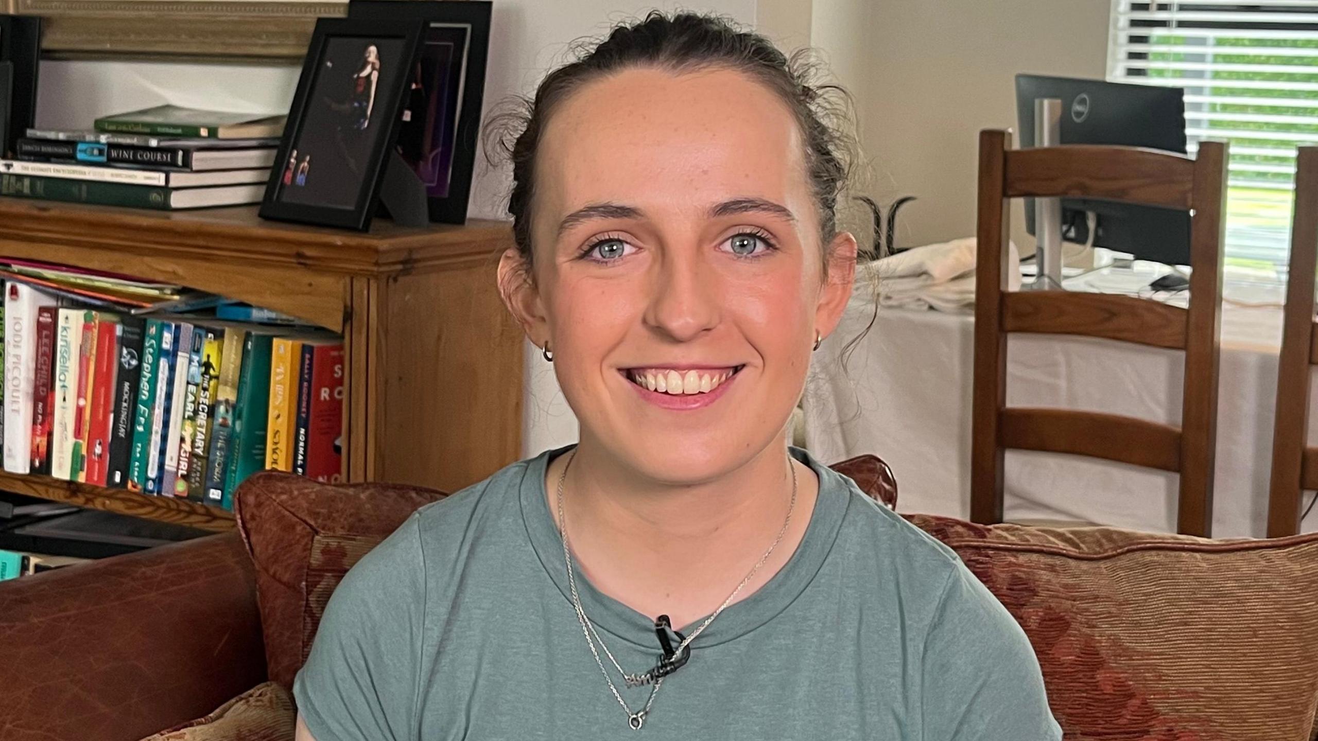 A young woman, Amy Symington, smiles broadly at the camera. She is sitting in the living room of a home. In the background, through an entrance into another room we can see a dining table. There is a also a low bookcase filled with books and topped with family photos. Amy has dark hair tied back and is wearing a light green t-shirt.