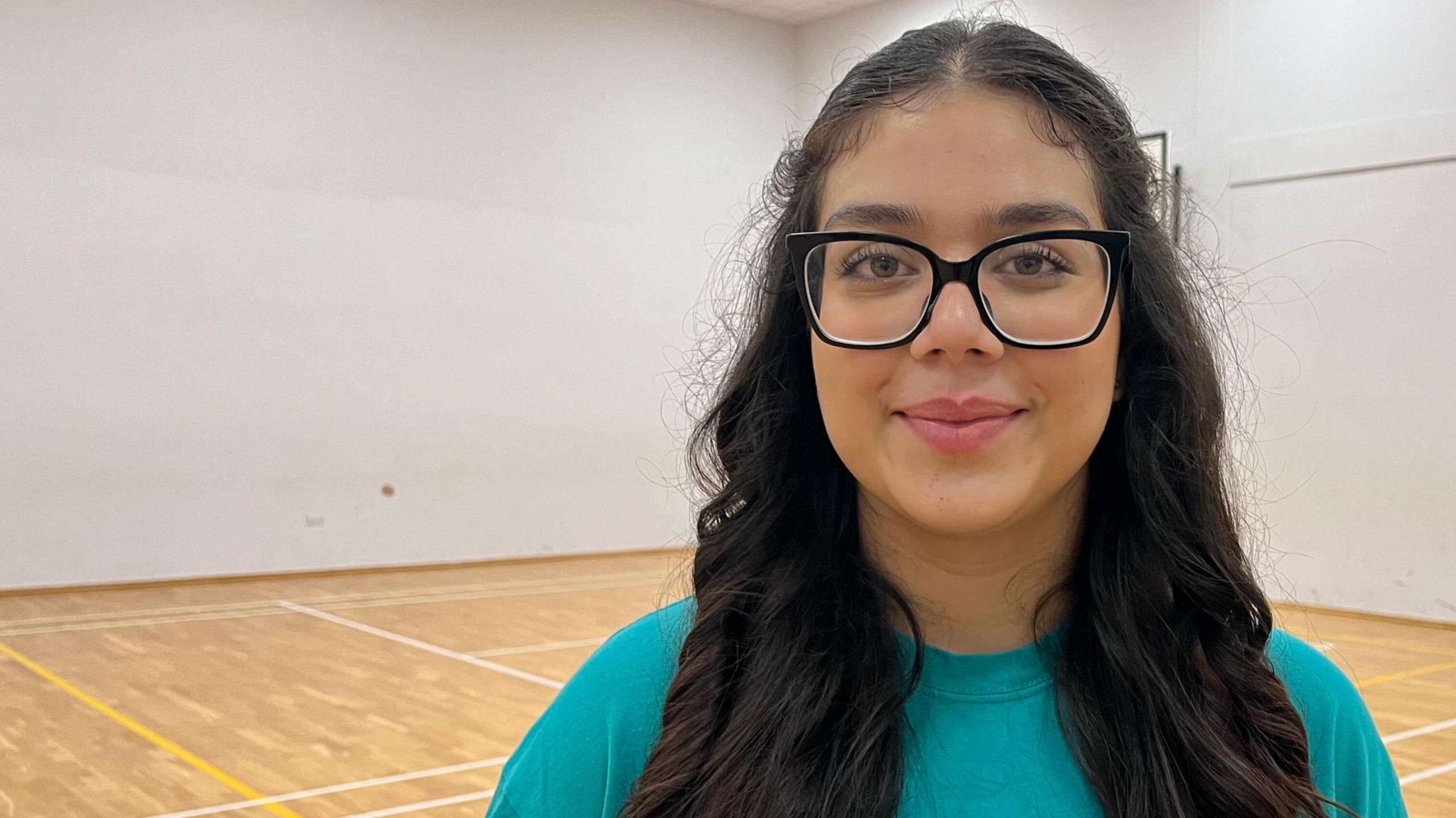 Hannah, a 15-year-old girl, stands in a white-walled school gym and looks into the camera. She has long black hair and is wearing an aquamarine T-shirt and large, black-rimmed glasses.