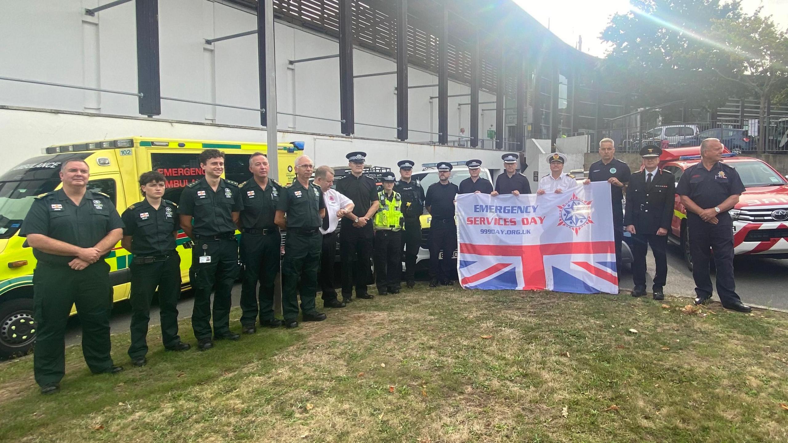 A group of emergency service personnel, including ambulance, fire, and police staff in their respective uniforms.  They are standing together outdoors in front of an ambulance, police car and a fire response vehicle. Several members of the group are holding a flag. The top half of the flag is coloured white with the words ‘Emergency Services Day’ printed on it. The lower half of the flag shows the bottom half of the Union Flag. 