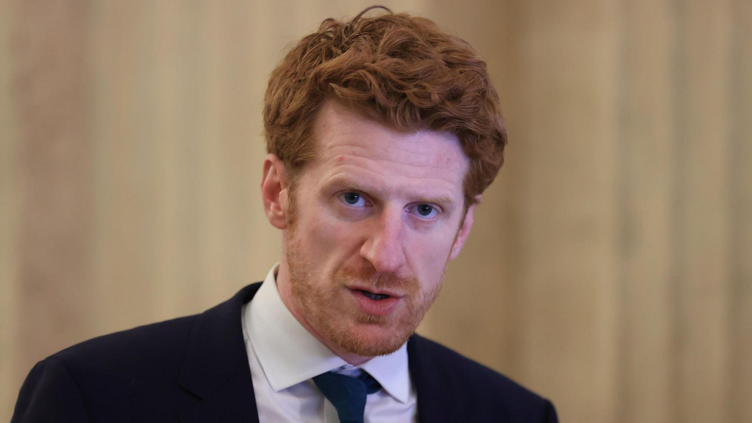 Matthew O'Toole  is wearing a suit with a white shirt and a tie. He has auburn hair. He's standing with white marble walls behind him.