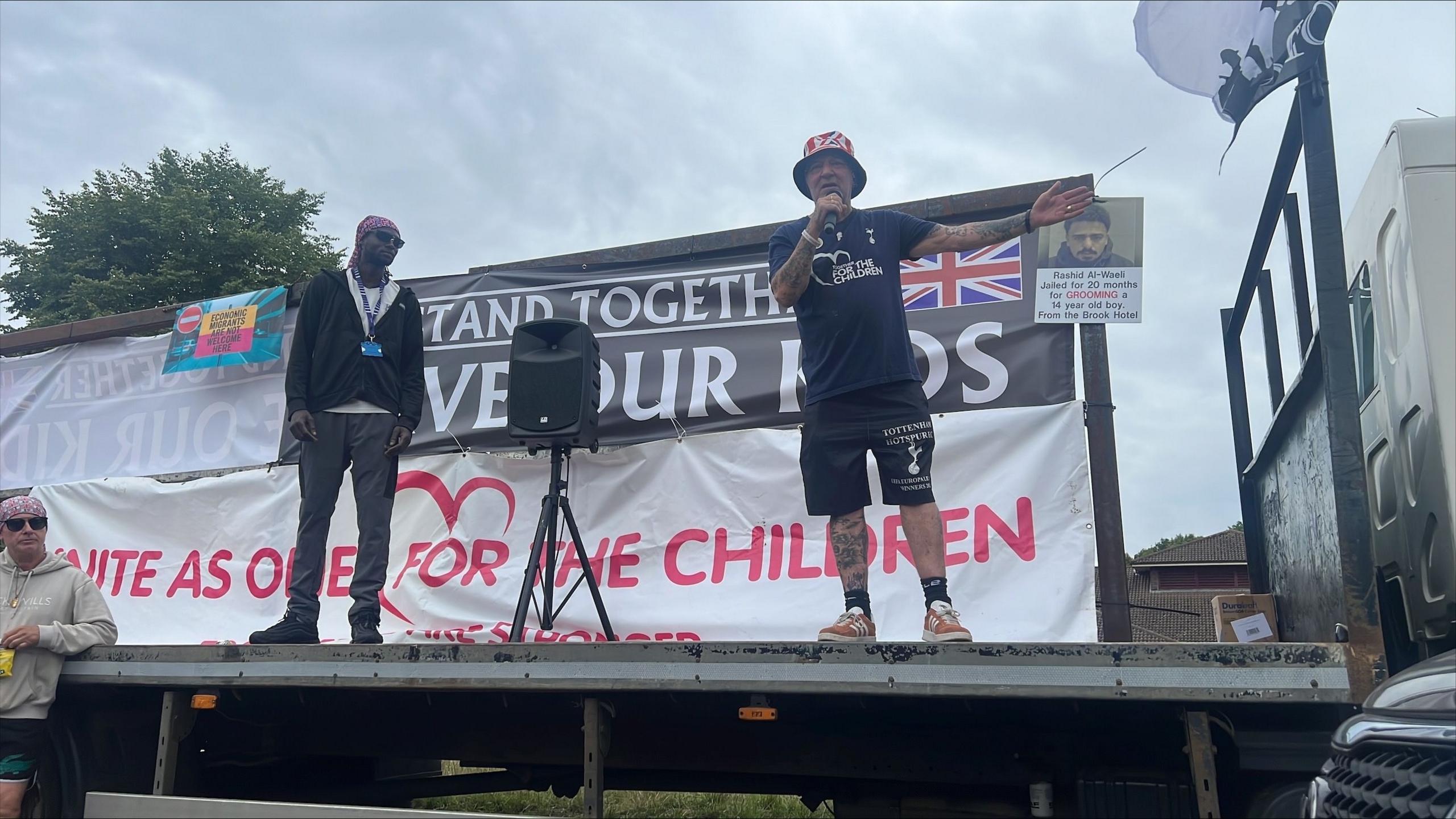 A man in black T-shirt and shorts and wearing a union jack bucket hat addresses a crowd from the back of a lorry. Another man stands beside him and looks on. Behind them are banners, one of which says "Unite as one for our children".