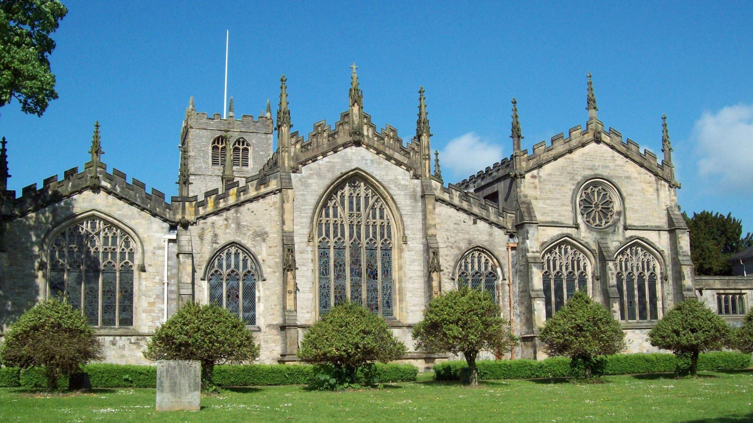 The Church of Holy Trinity with several large stained glass windows. It is a stone-coloured and a large impressive building next to trees.