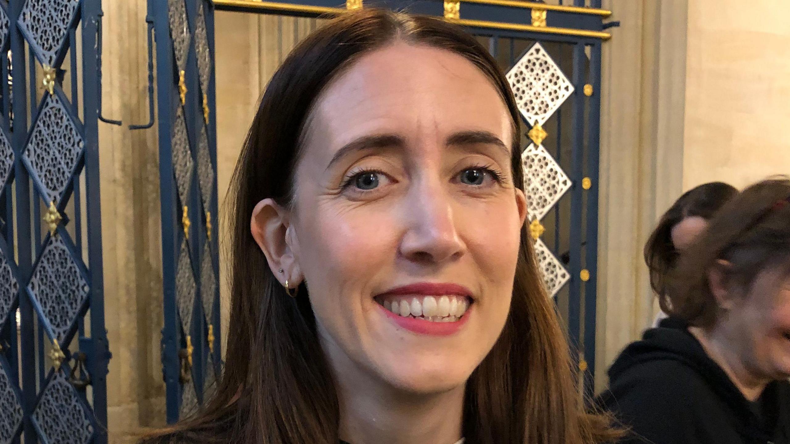 Catherine Rayson with shoulder length dark hair, smiling at the camera, standing in St Edmundsbury Cathedral 