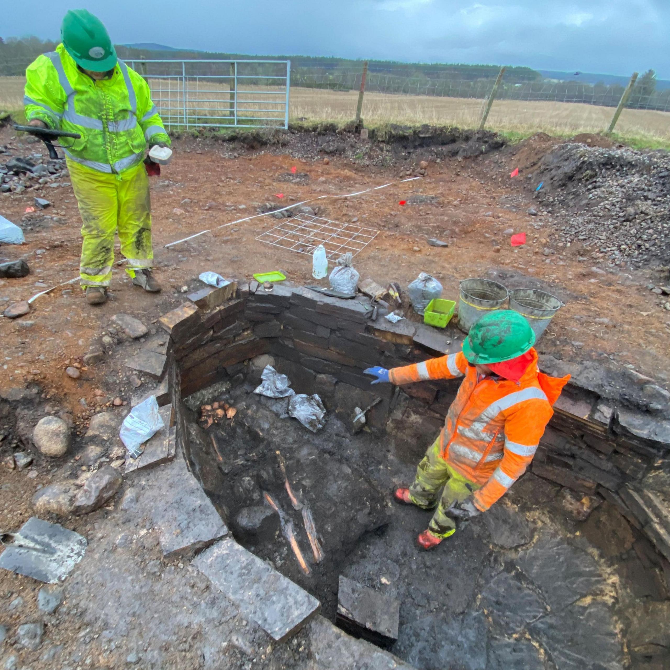 Two people wearing green hard hats and high visibility clothing stand at the stone-lined burial pit. The scene is set within a rural landscape of fields and trees.