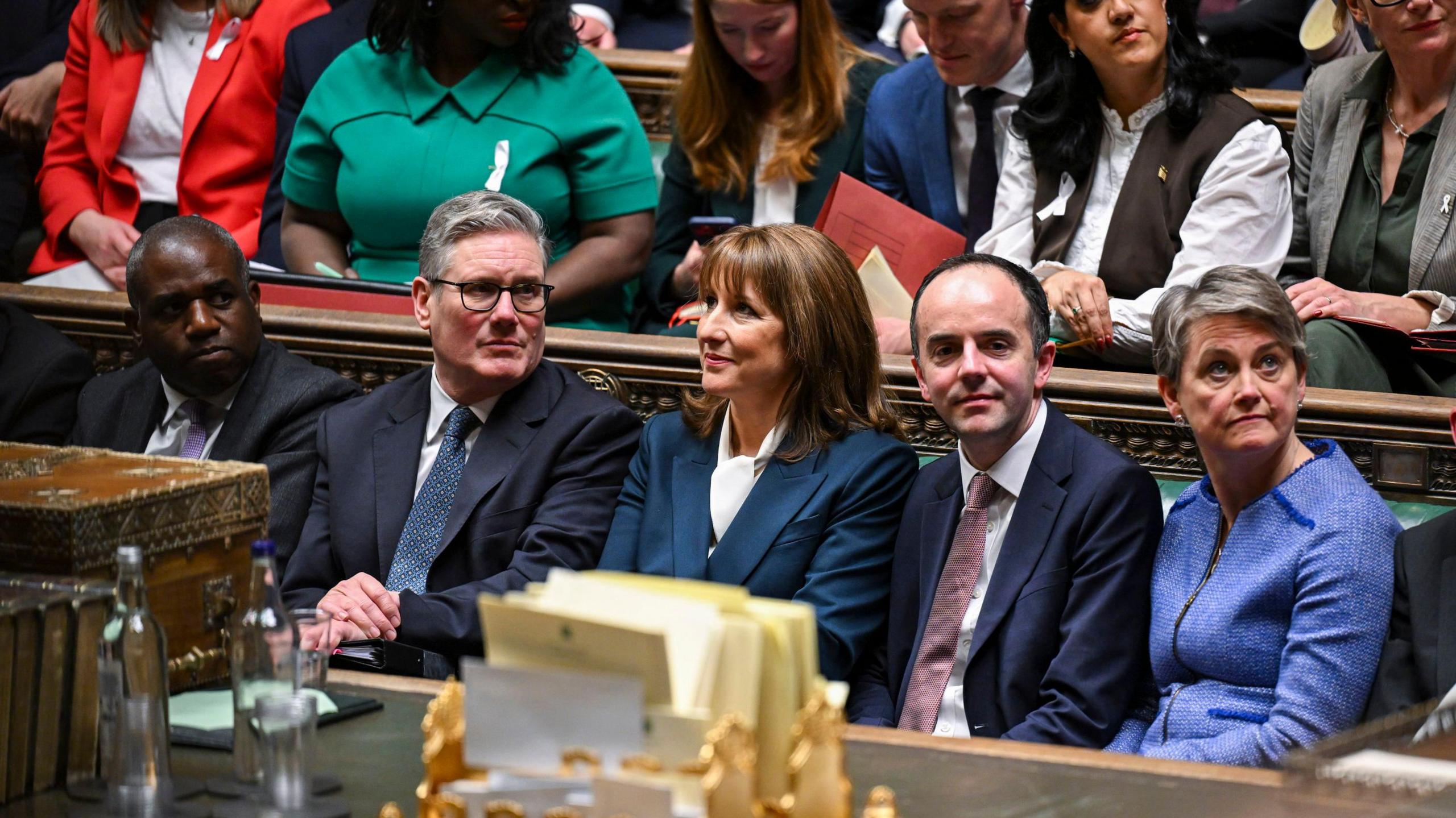 The Chancellor Rachel Reeves is pictured inside the House of Commons alongside the Prime Minister and other MPs. She is sat on the green benches, looking across the room.