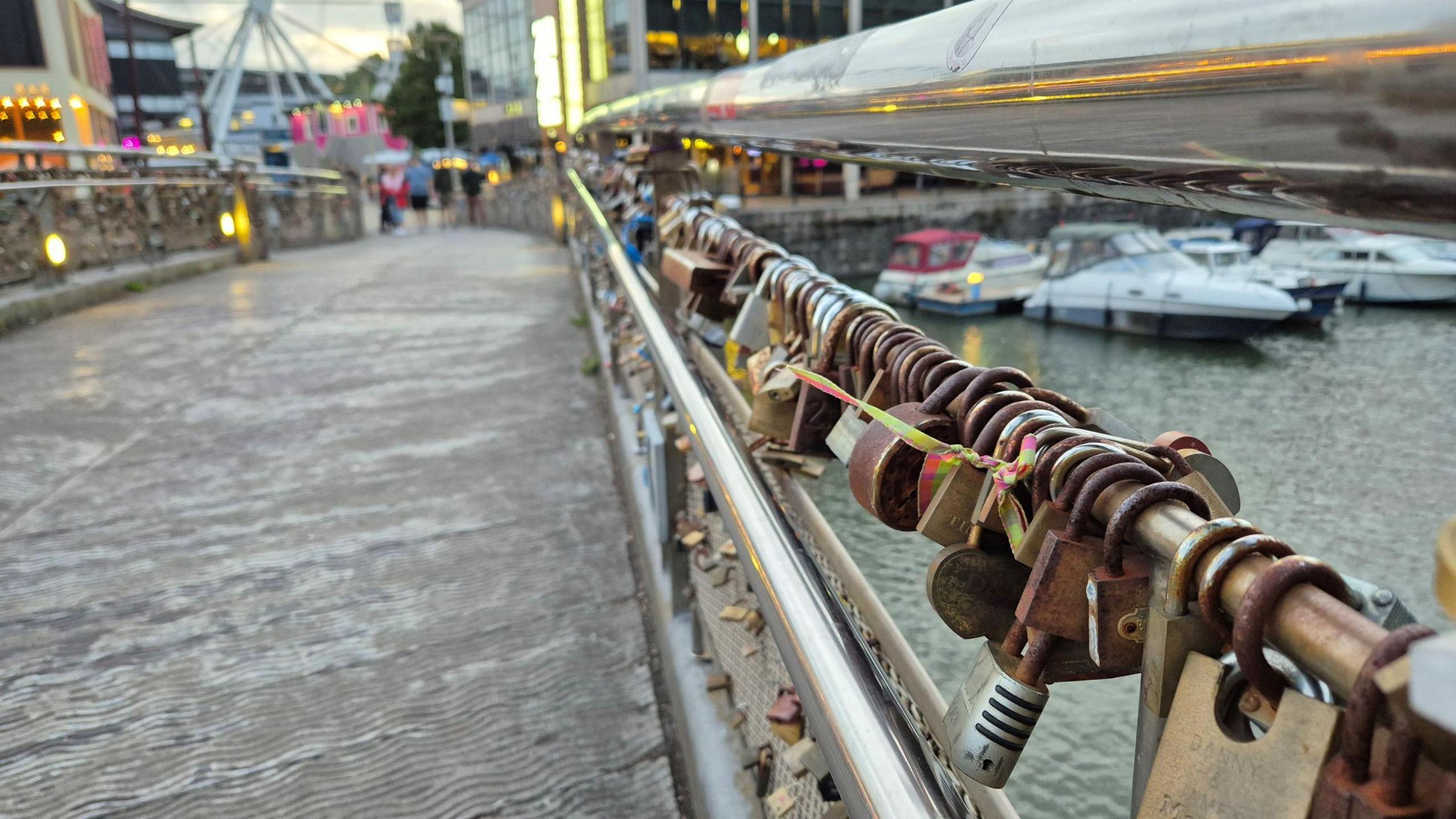 A long line of padlocks is seen close up on Pero's Bridge in Bristol city centre. In the background, but not in focus, are some small boats.