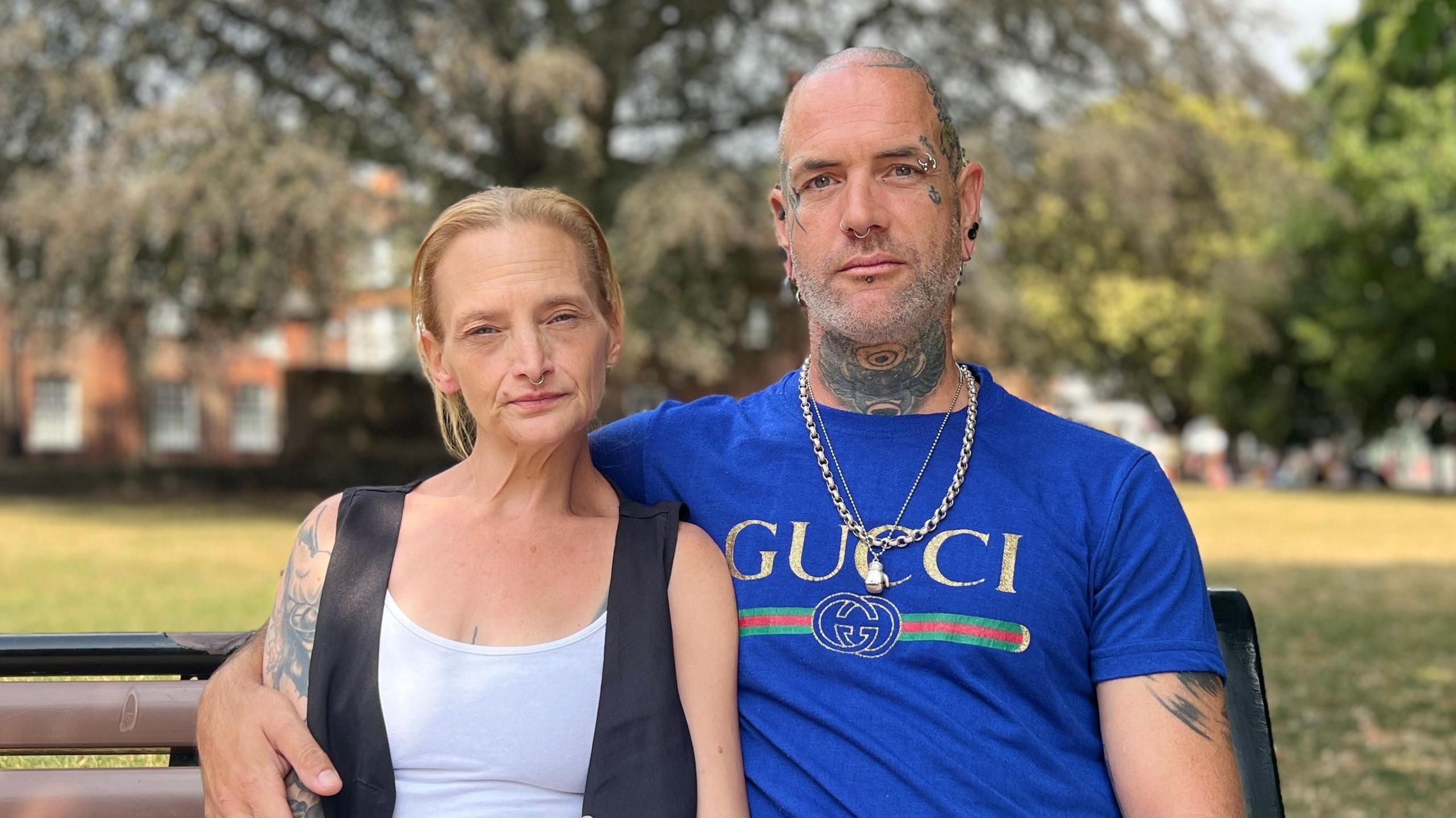 Rebecca and Tony Nye are sitting on a bench with trees and grass visible but not in focus behing. Tony has his arm around Rebecca and is wearing a blue "Gucci" shirt with gold chains around his neck. He has partial tattoos visible on his arm, head and neck and has facial piercings. Rebecca is wearing a white top with balck waistcoat over and has a tattoo on her arm. Both are looking at the camera with neutral expressions.