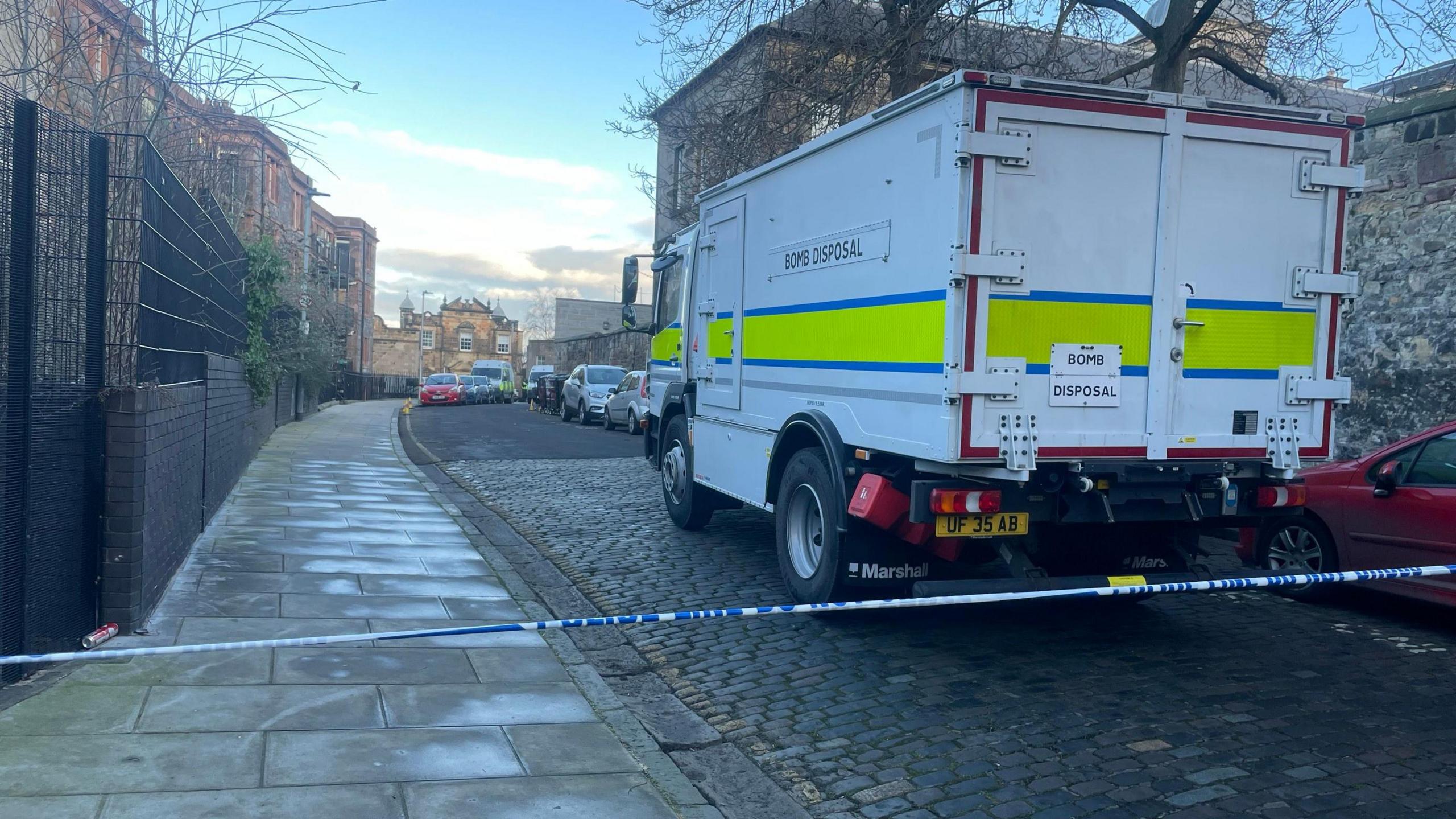 A bomb disposal van parked on High School Yards in Edinburgh. The van is white, with a yellow and blue stripe round it. It has the words Bomb Disposal in black lettering on the side. A thin line of white and blue police tape is in the foreground.