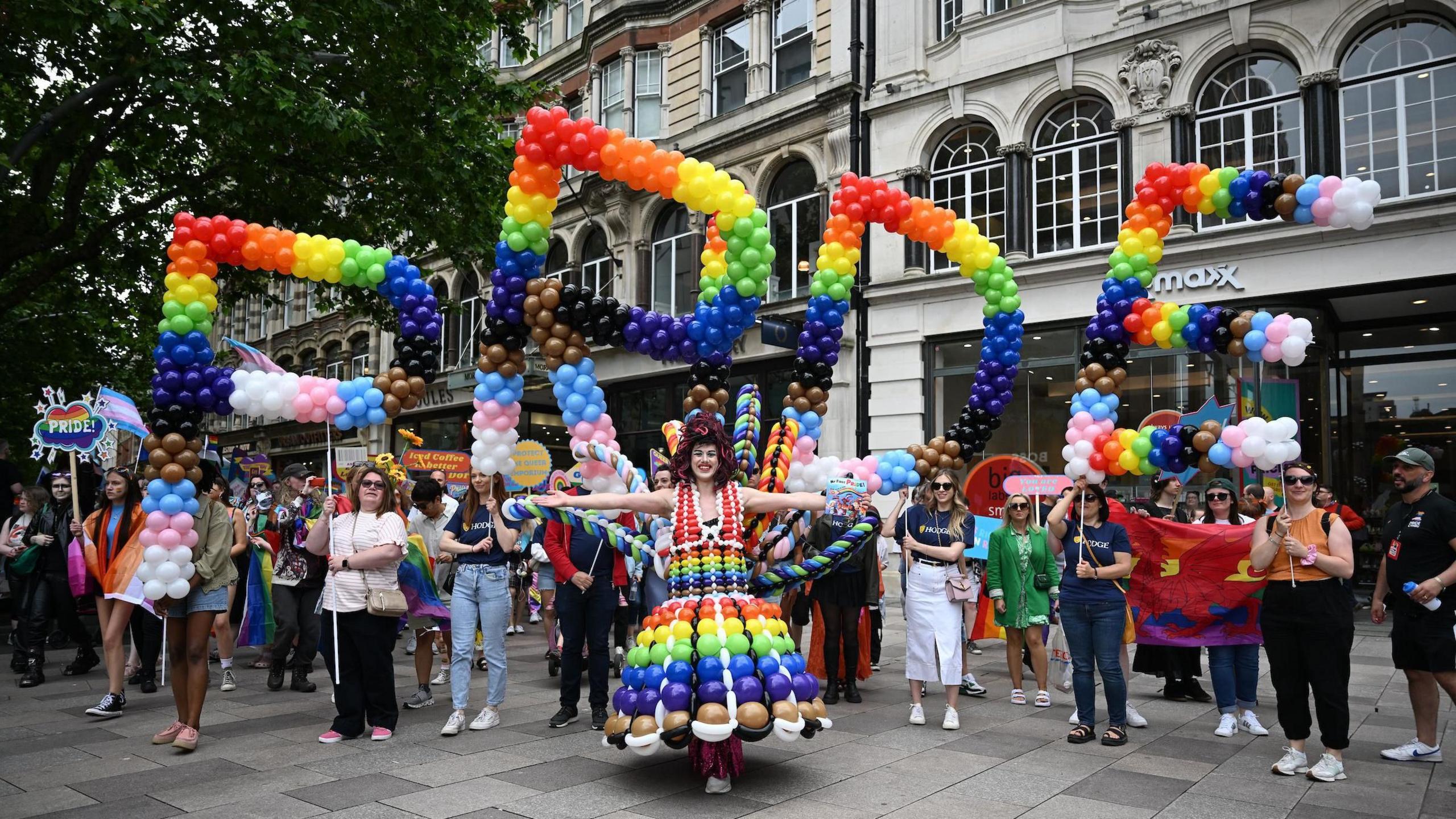 A person wearing a colourful balloon outfit, behind them is a colourful rainbow balloon sign that says Pride, with loads of people behind them.