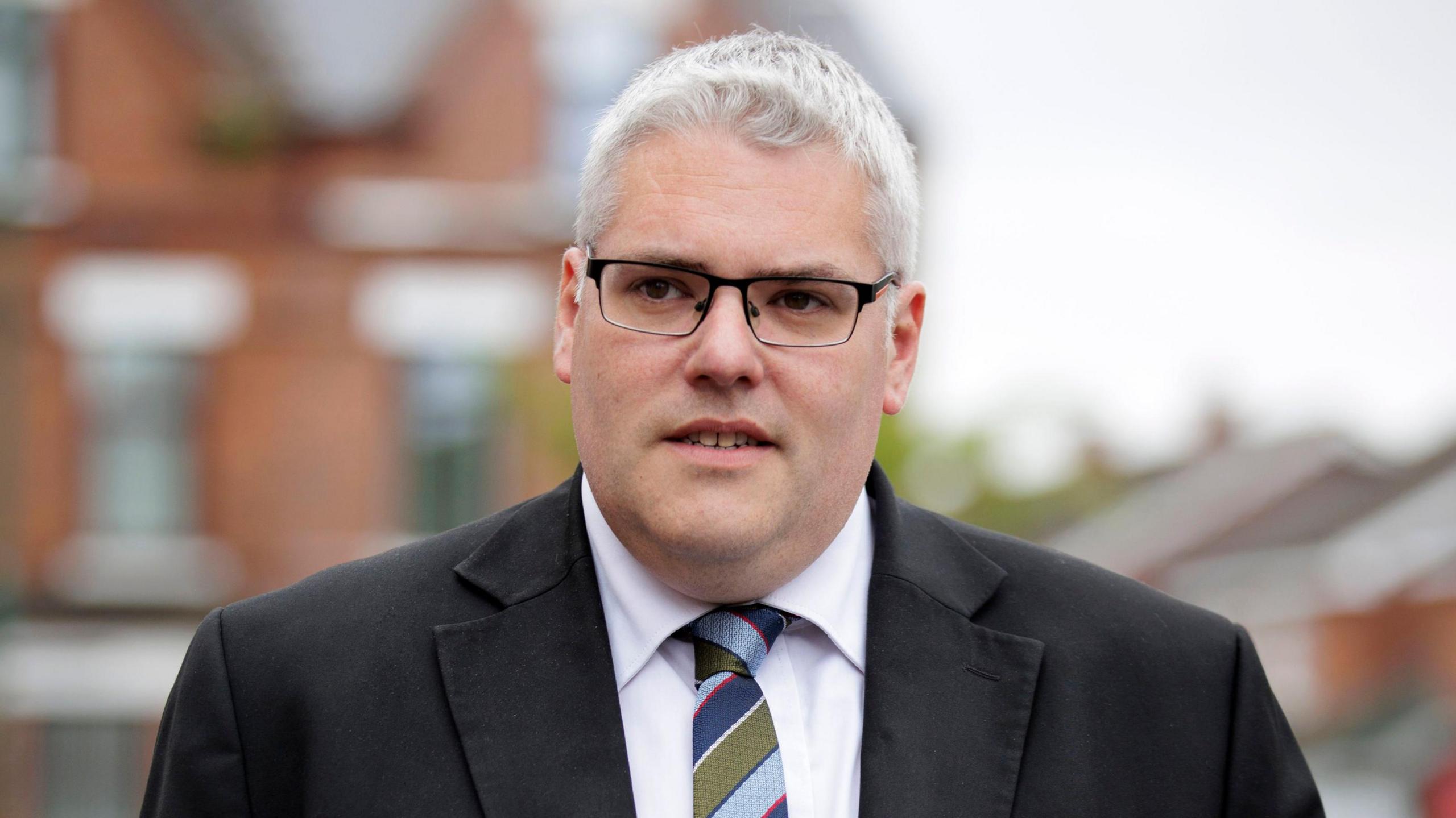 A picture of Gavin Robinson, who has short, white hair and is wearing slim framed black glasses. He is also wearing a black suit with a white shirt and a light blue, navy, olive green and red lined tie. He is standing in front of red brick houses.