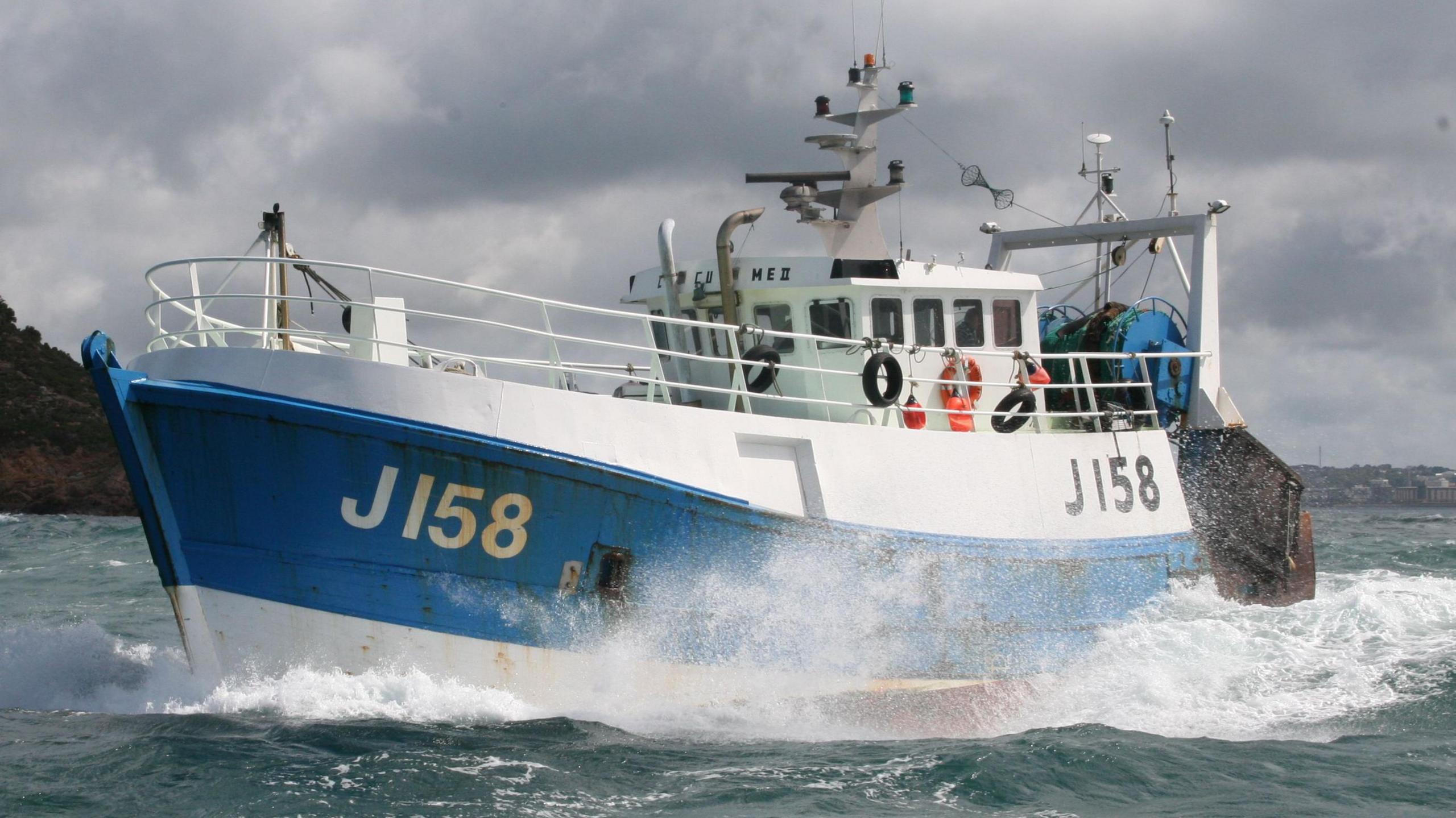 A white and blue fishing boat with J158 on the left side, in the ocean, next to another vessel out at sea with grey skies and land seen in the background.
