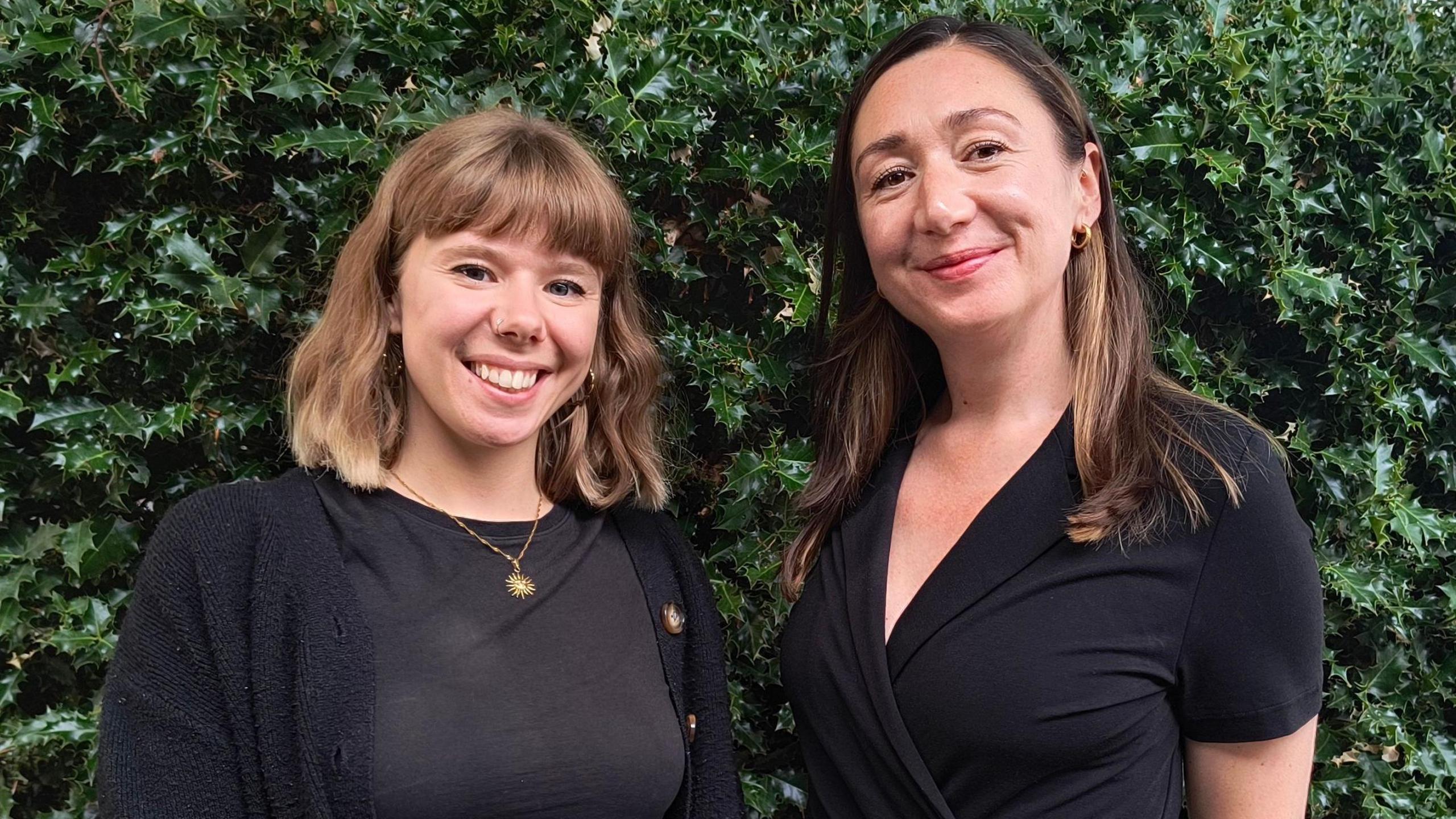 Two women standing in front of a short wall and a holly bush. Both are wearing dark clothing, have brown hair and are smiling.