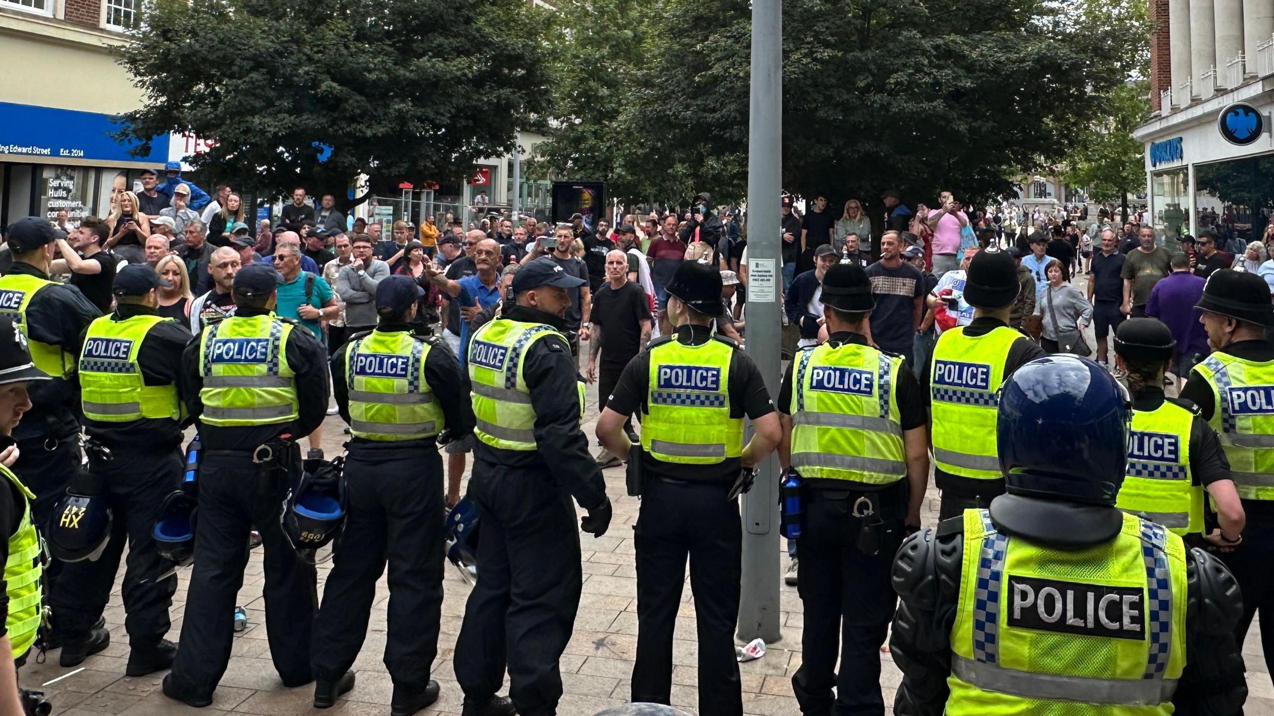 Police officers lined up in a row in Queen Victoria Square, some with riot helmets and standing in front of a group of people