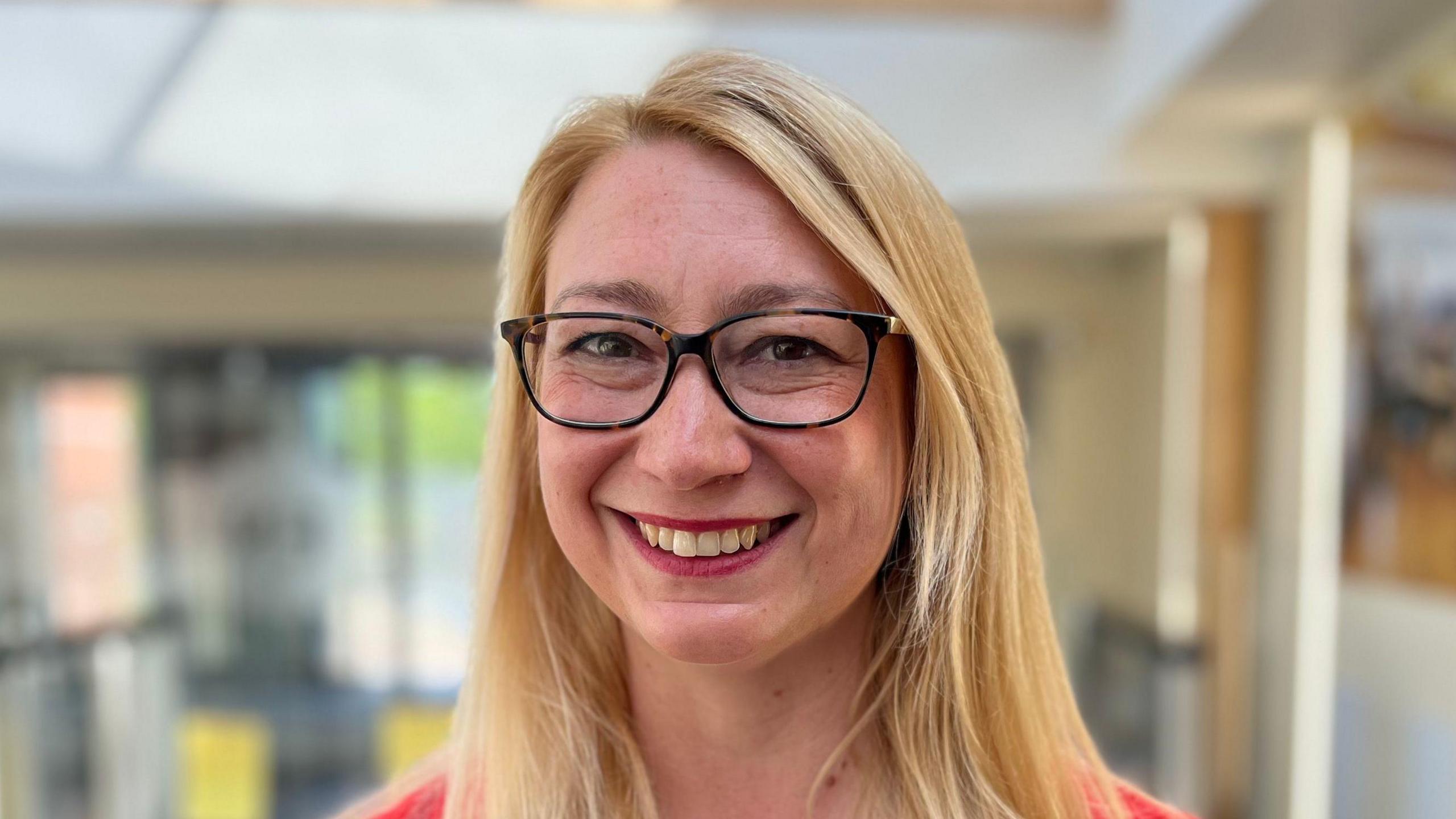 Jess Asato smiles at the camera. She has long blonde hair and wears black glasses.