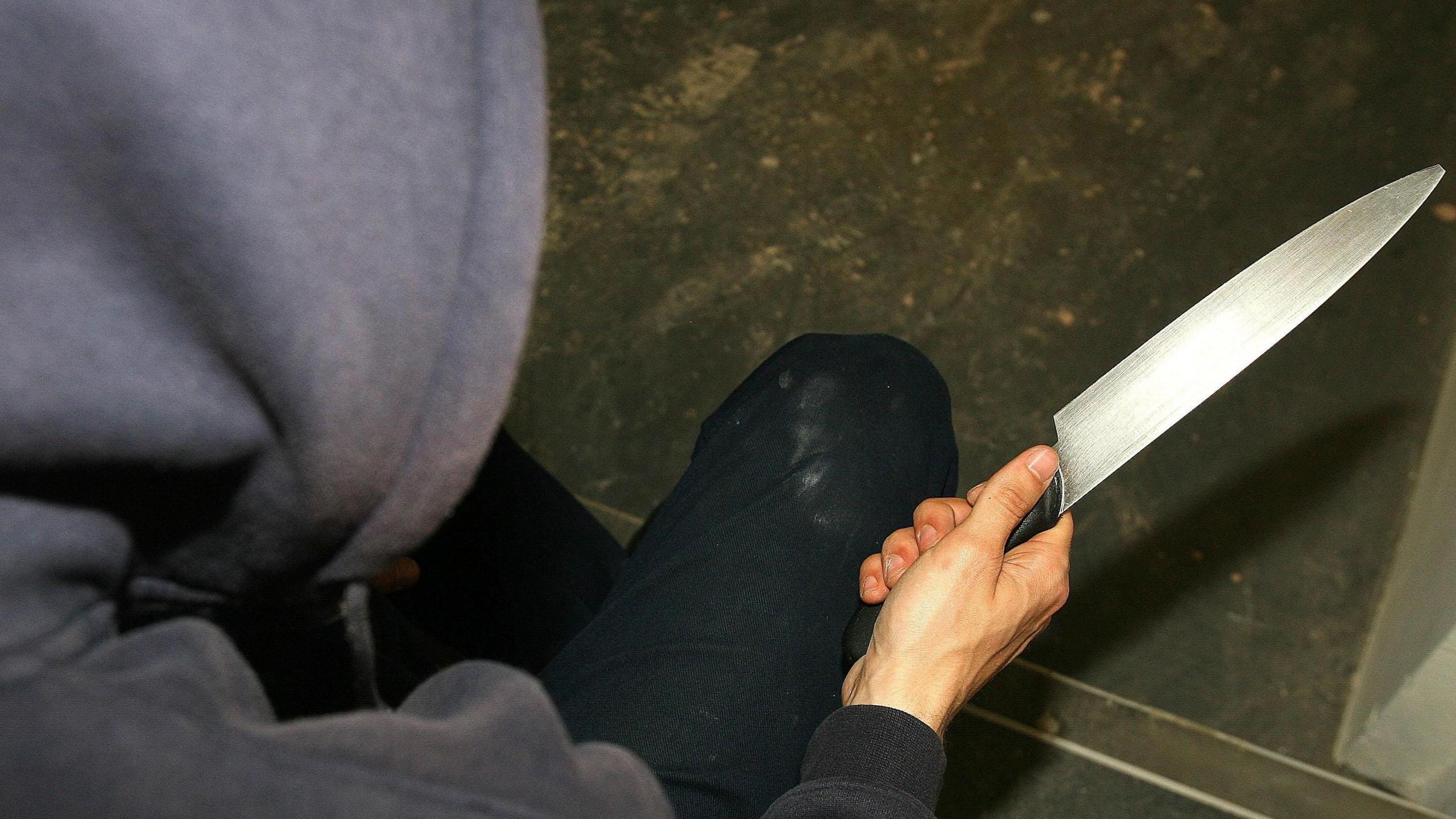 A generic photo of a man in a blue hoodie holding a large knife. The photo is taken from above the man and looking down toward his hand holding the knife.