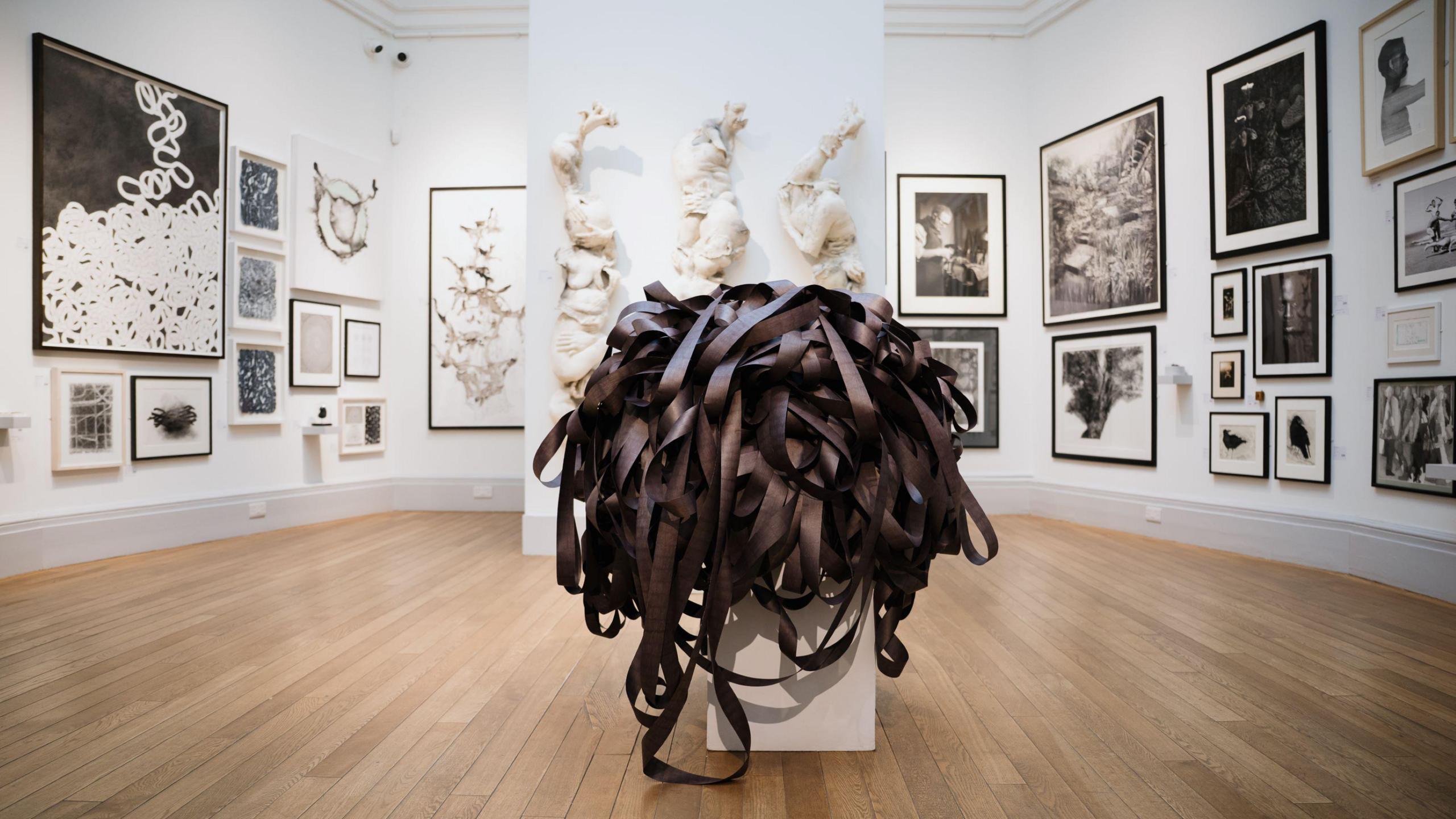 An art gallery with dozens of paintings on its walls and in the foreground on a small plinth is a collection of what appear to be dark ribbons meshed together in piles. The floor of the gallery - the RWA in Bristol - is of light wooden boards.