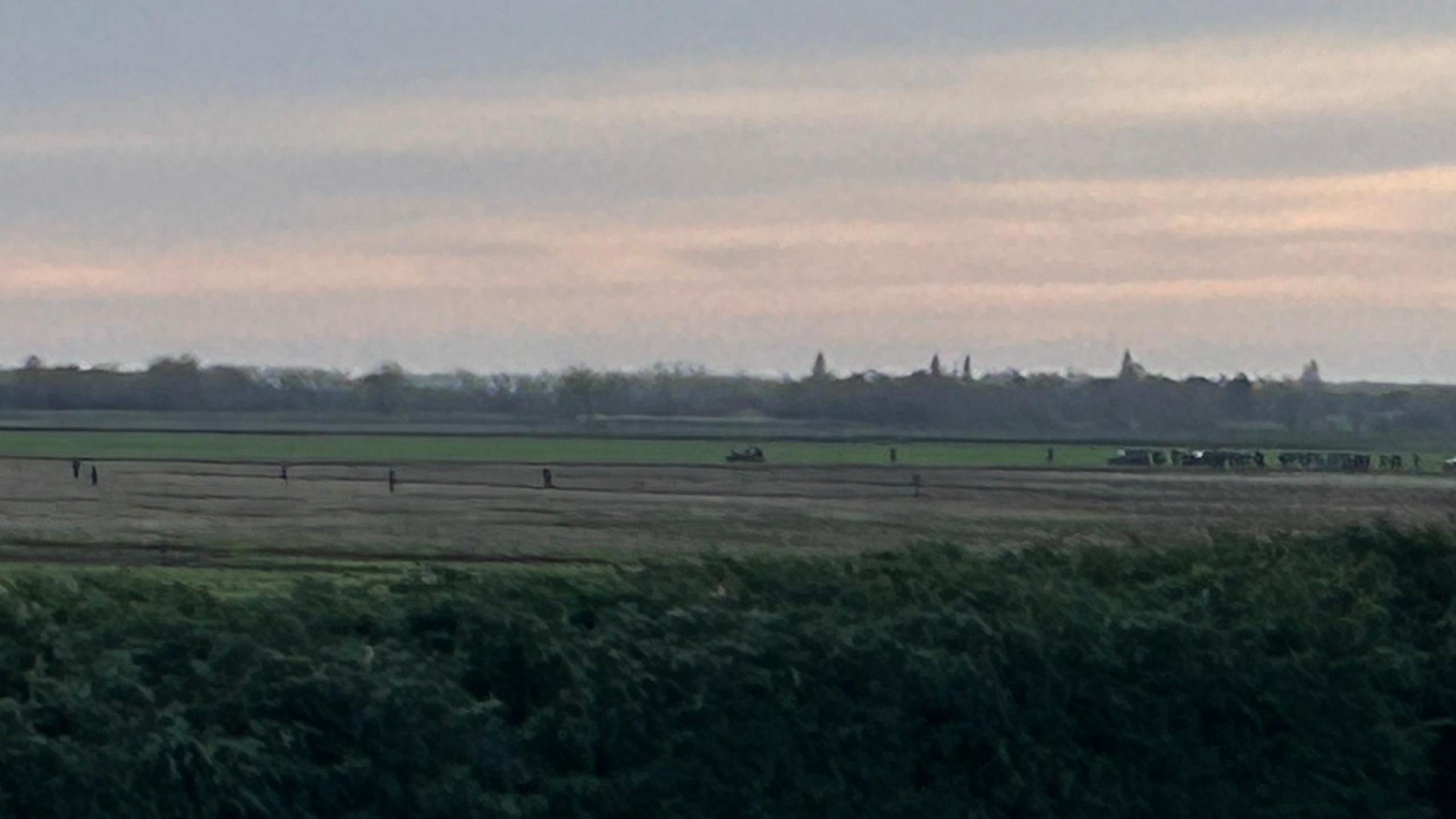 From a distance, a gang of hare coursers can be seen walking and driving through a field. The sun is setting and it is getting dark. 