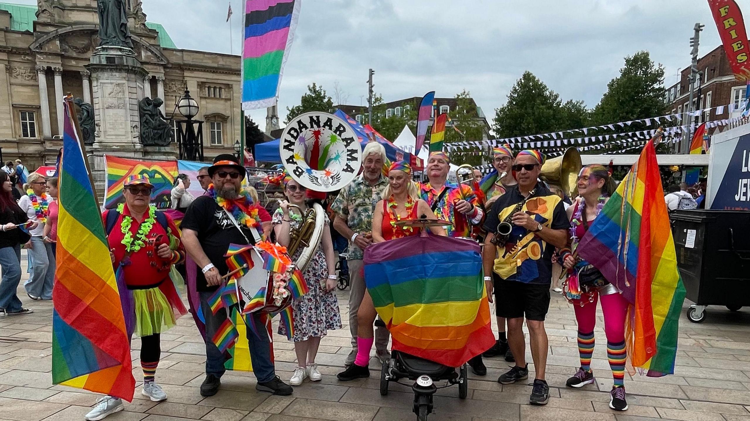 A group of people carrying rainbow flags and wearing colourful clothes.  A woman in the middle has a tuba, with the "Bandanarama" logo on it.