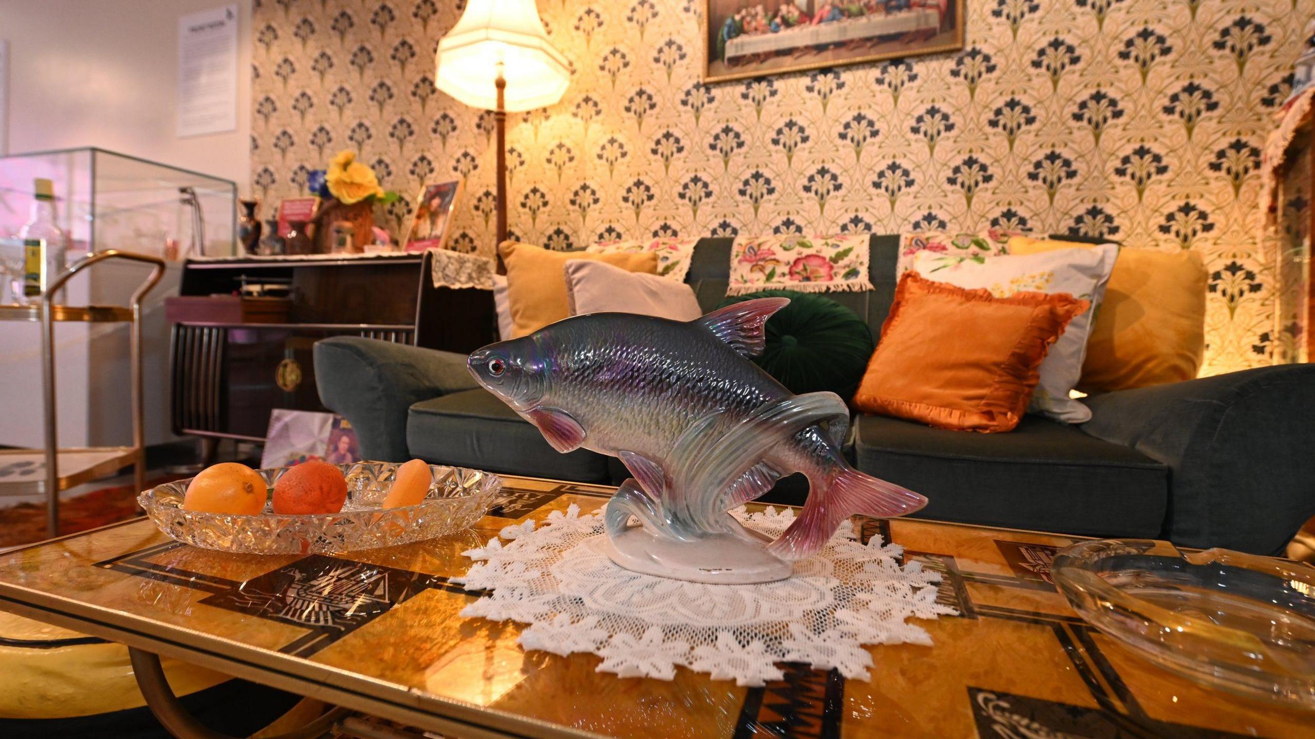 The front room featured in the exhibition. There is a coffee table in the foreground with a glass fish ornament on top and a glass fruit bowl with two oranges and a banana. In the back there is a green sofa with orange, white and yellow cushions and white and blue wall paper behind it.