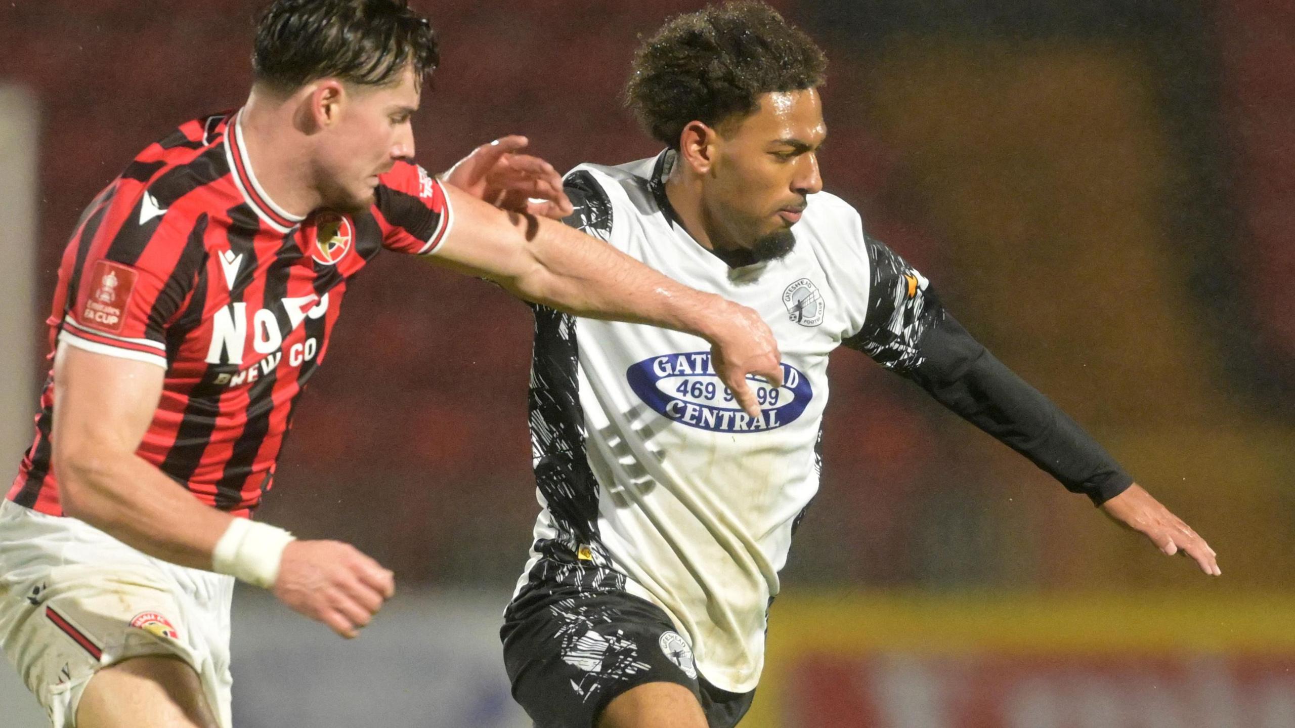 Tyrelle Newton in white Gateshead shirt, with black undershirt sleeves and shorts, battling with a Walsall player on the left with red and black stiped shirt