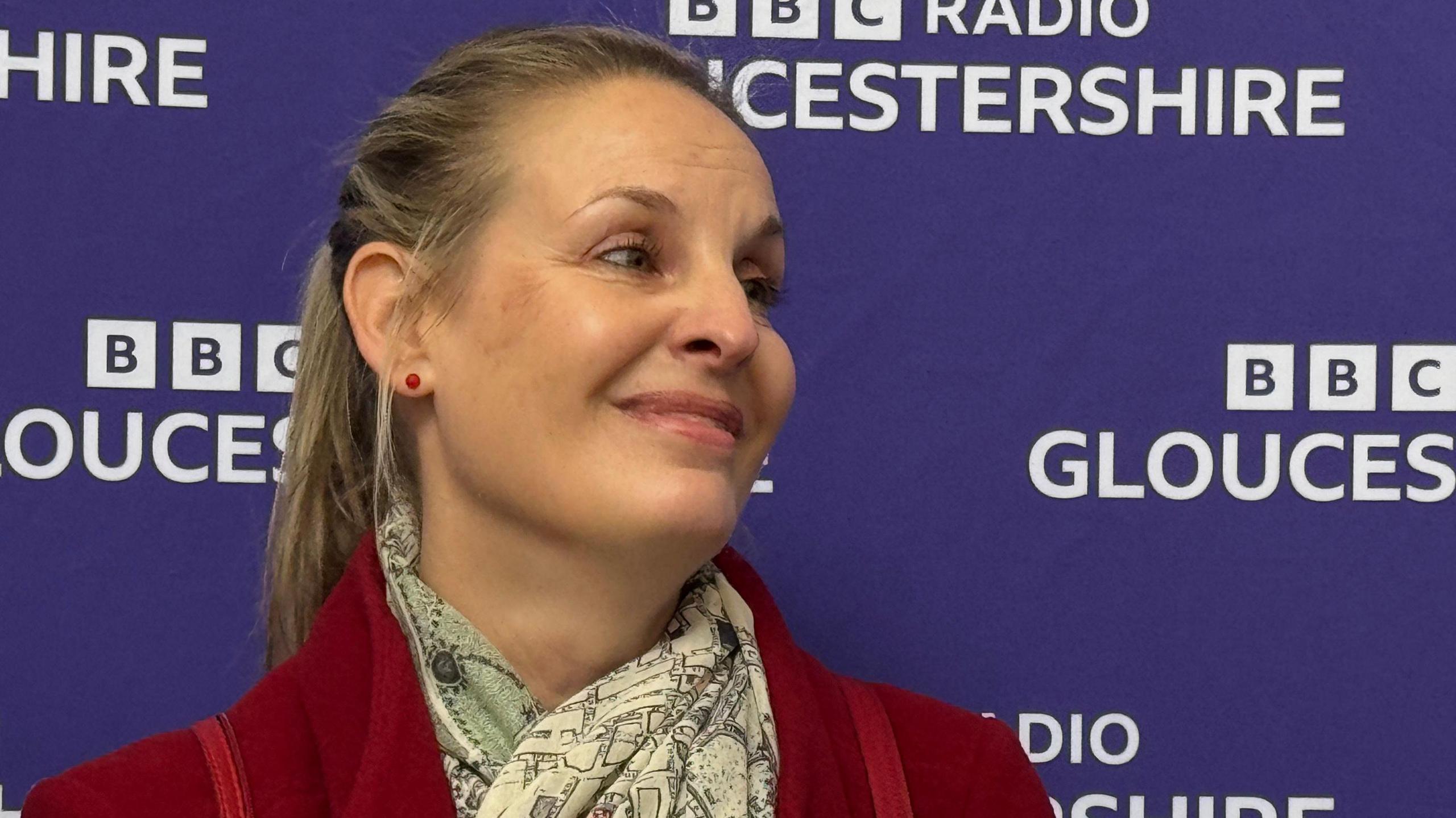 A woman with long blonde hair in a ponytail who is wearing red stud earrings, a red blazer, and a white fashion scarf with a map pattern on it. She is smiling and looking up and is stood against a purple background that reads "BBC Radio Gloucestershire".