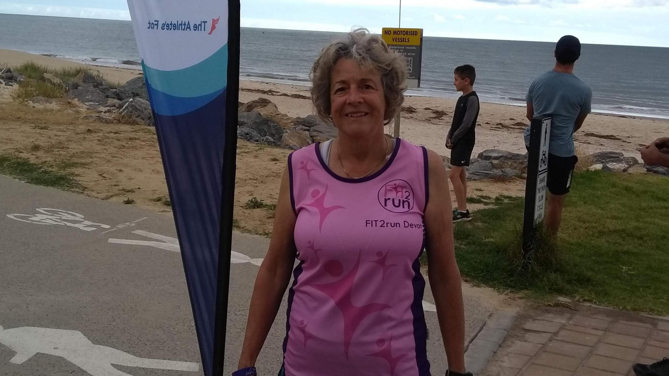 Faye Hussey smiling at the camera. She is wearing a pink top and is stood in front of a sandy beach. 