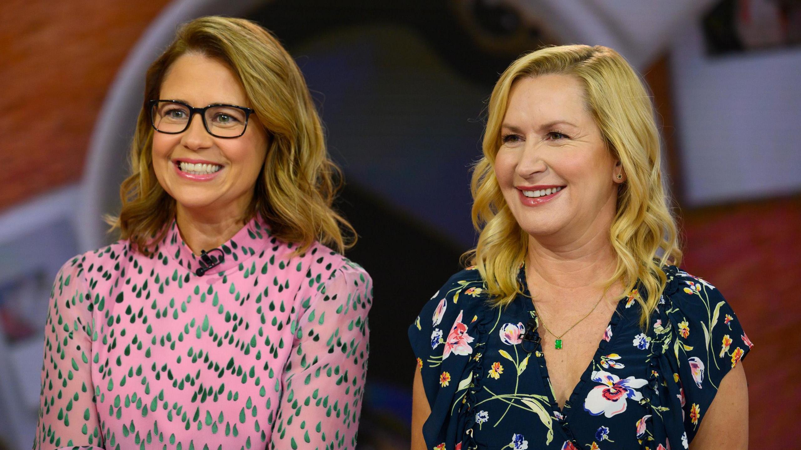 Jenna Fischer and Angela Kinsey pictured in 2022. Jenna has curled dark blonde hair and wears brown rectangle-rimmed glasses and a pink high-necked blouse. Angela, who has shoulder-length blonde hair, wears a dark blue shirt with a floral pattern. 