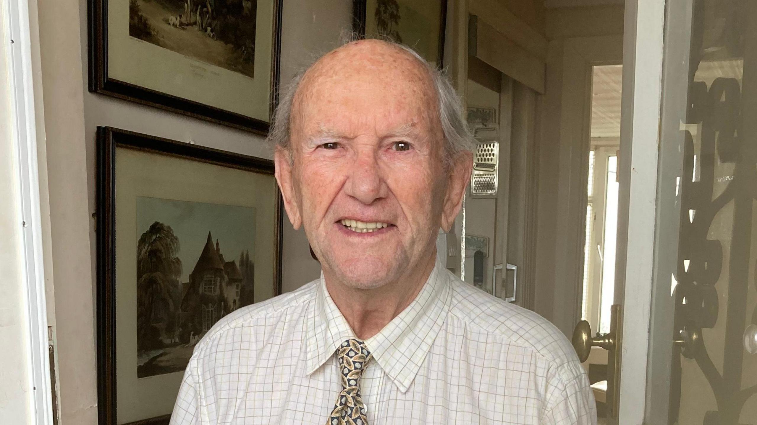 Tony Bird smiling at the camera. He is standing at the doorway of a house with pictures behind him.