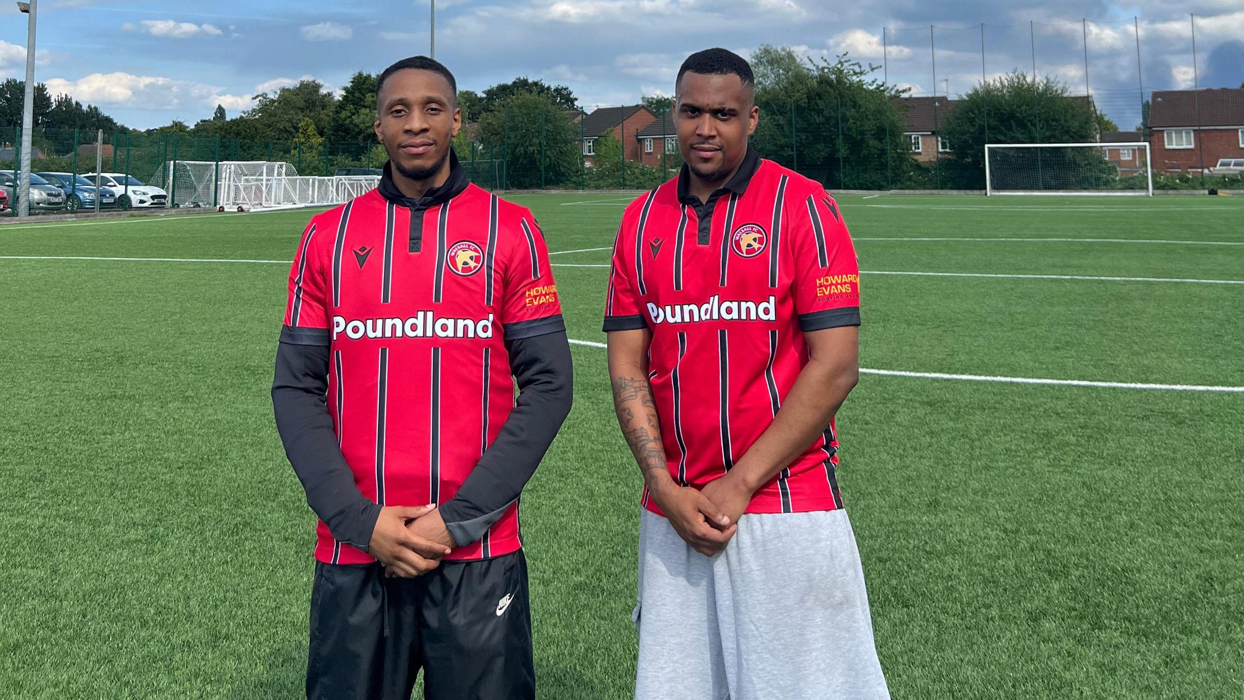 Two men wearing Walsall kit stand on a football pitch with a goal behind them