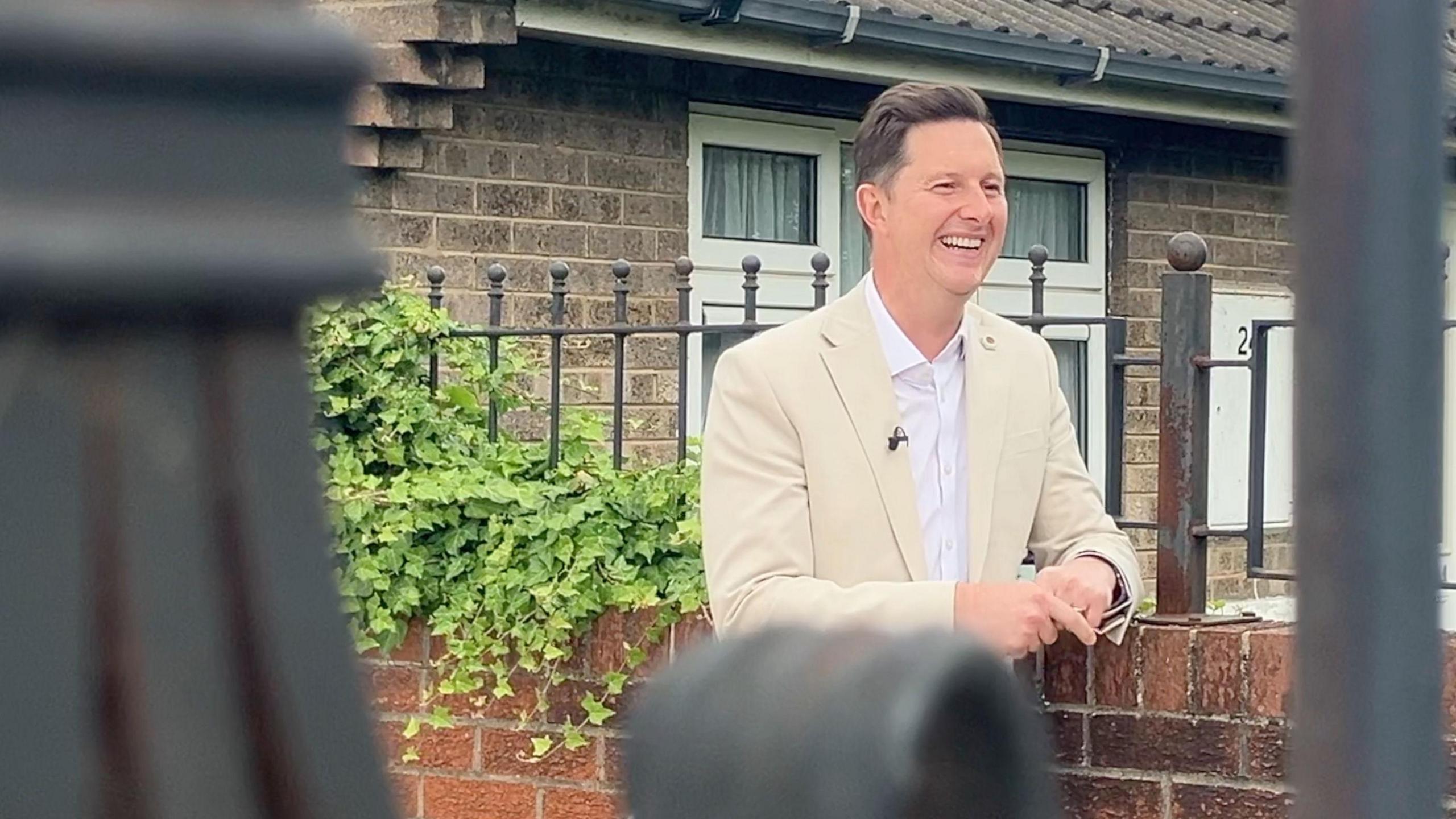Doncaster East and Isle of Axholme MP Lee Pitcher smiles as he leans on a low red brick wall. He is wearing a cream suit jacket with a white shirt.